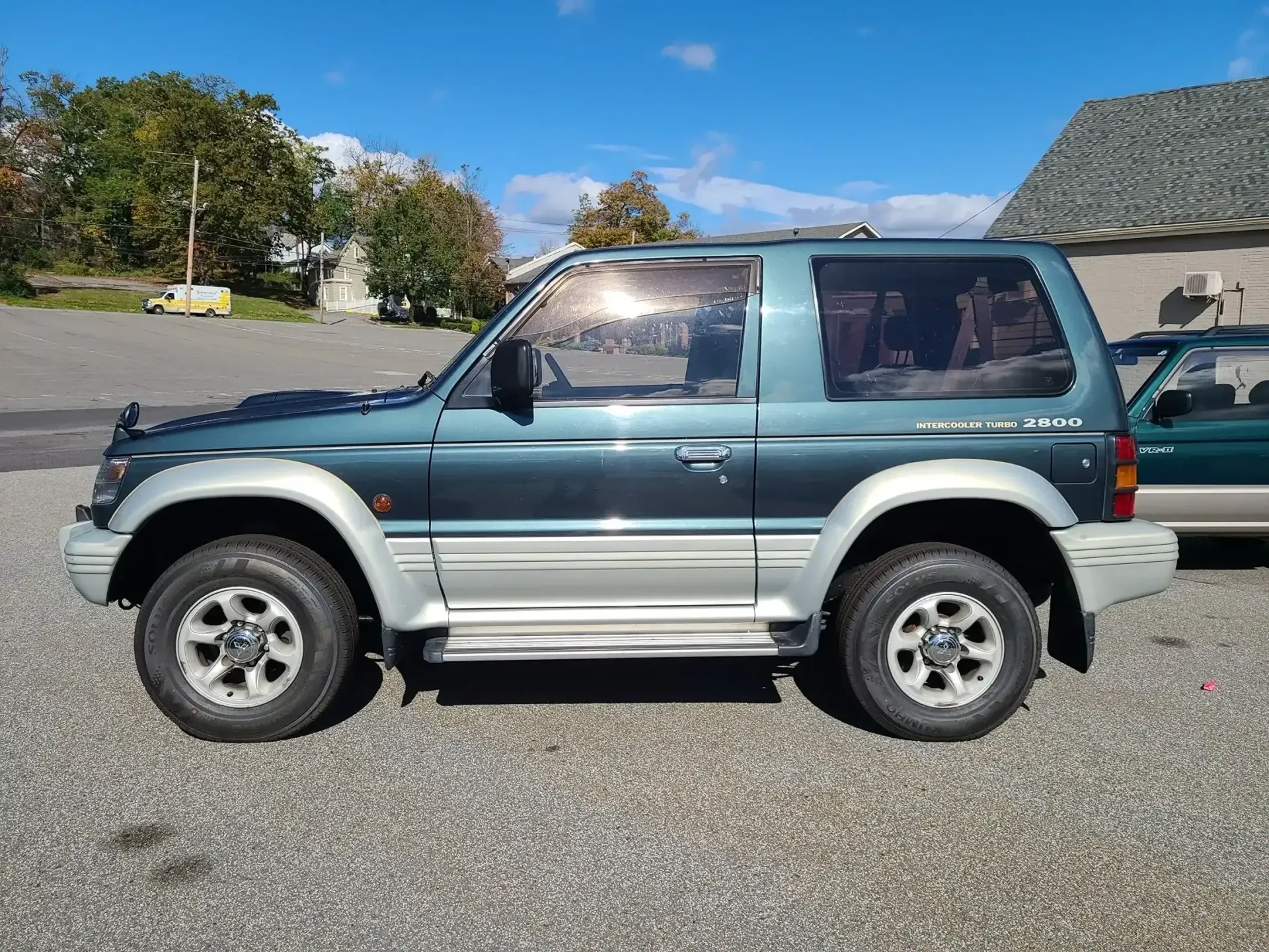 A two-door, dark teal and silver Mitsubishi Pajero SUV parked on a gravel lot under a clear blue sky.
