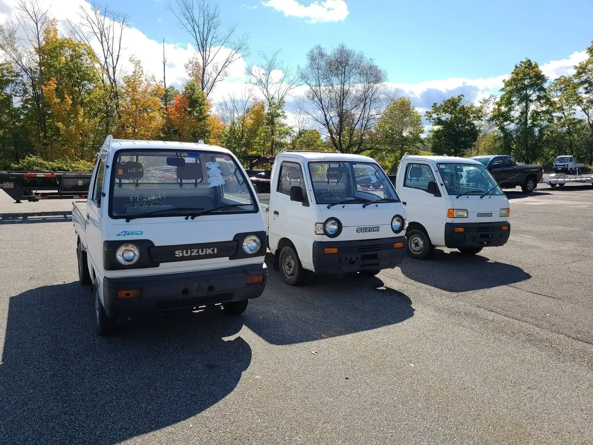 Three white compact pickup trucks parked on a gravel lot under a bright, sunny sky with trees in the background.