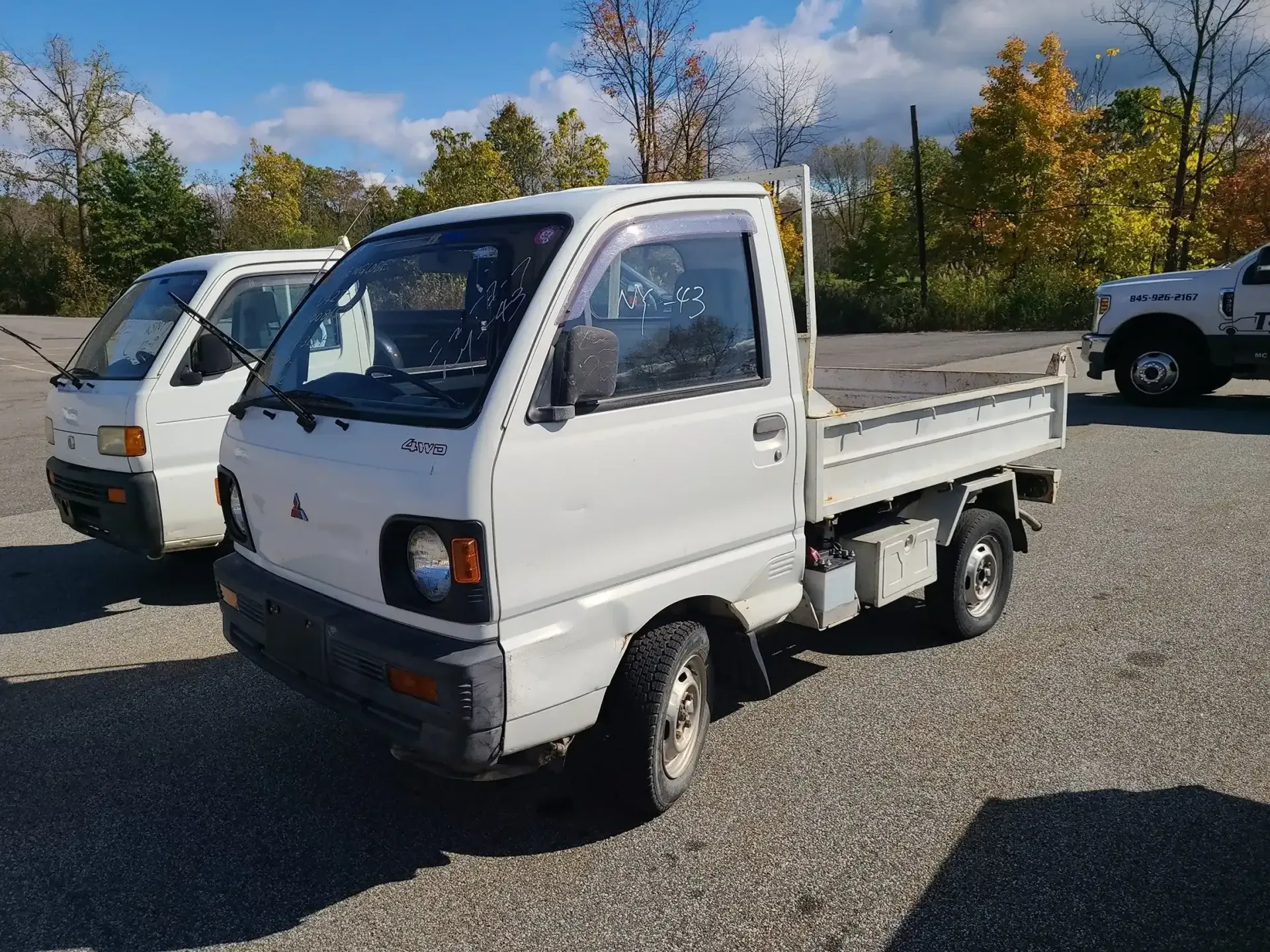 Two white Kei trucks parked on a gravel lot under a sunny, blue sky.