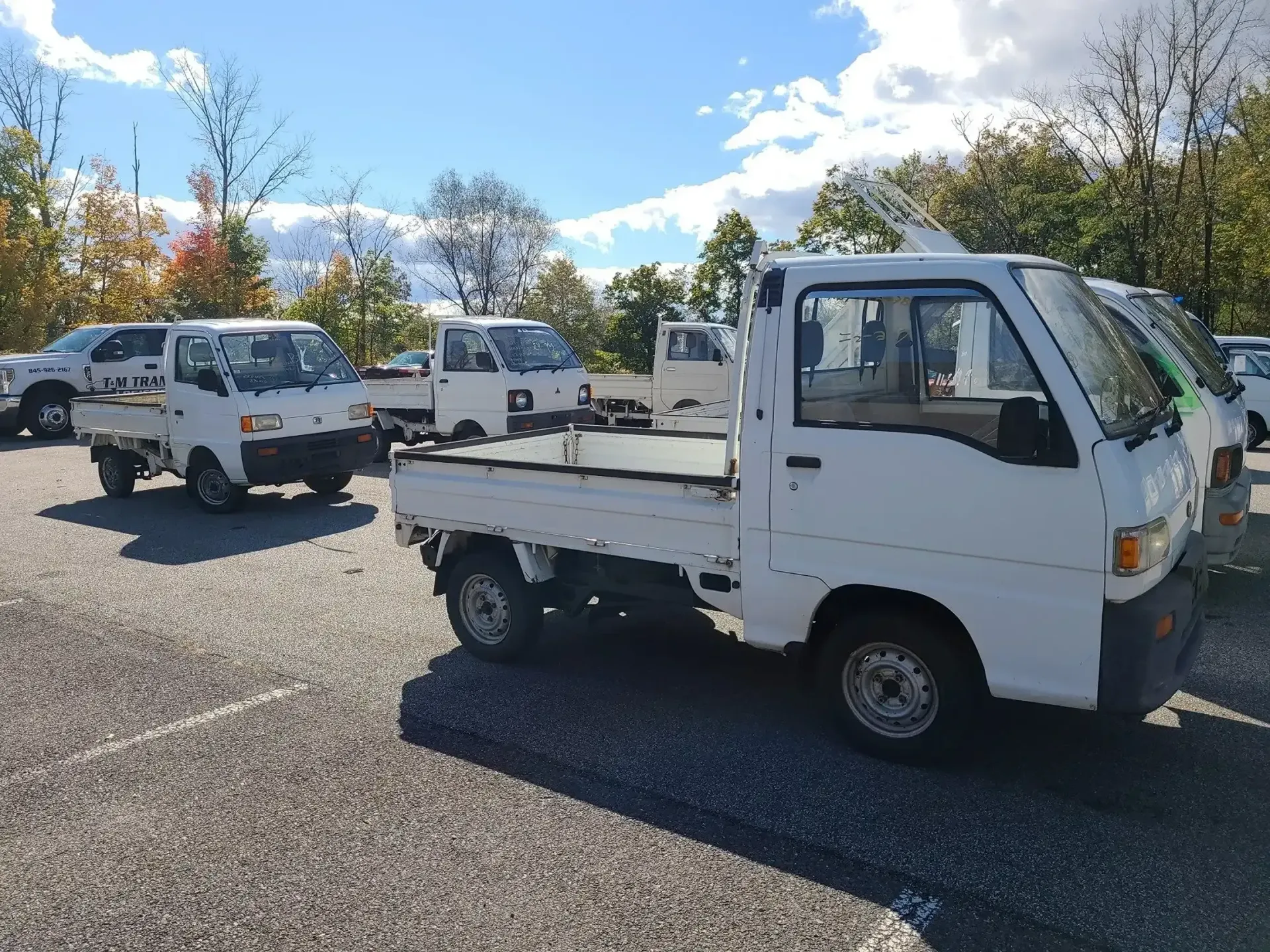 A fleet of white mini-trucks parked on a gravel lot under a bright, partly cloudy sky.
