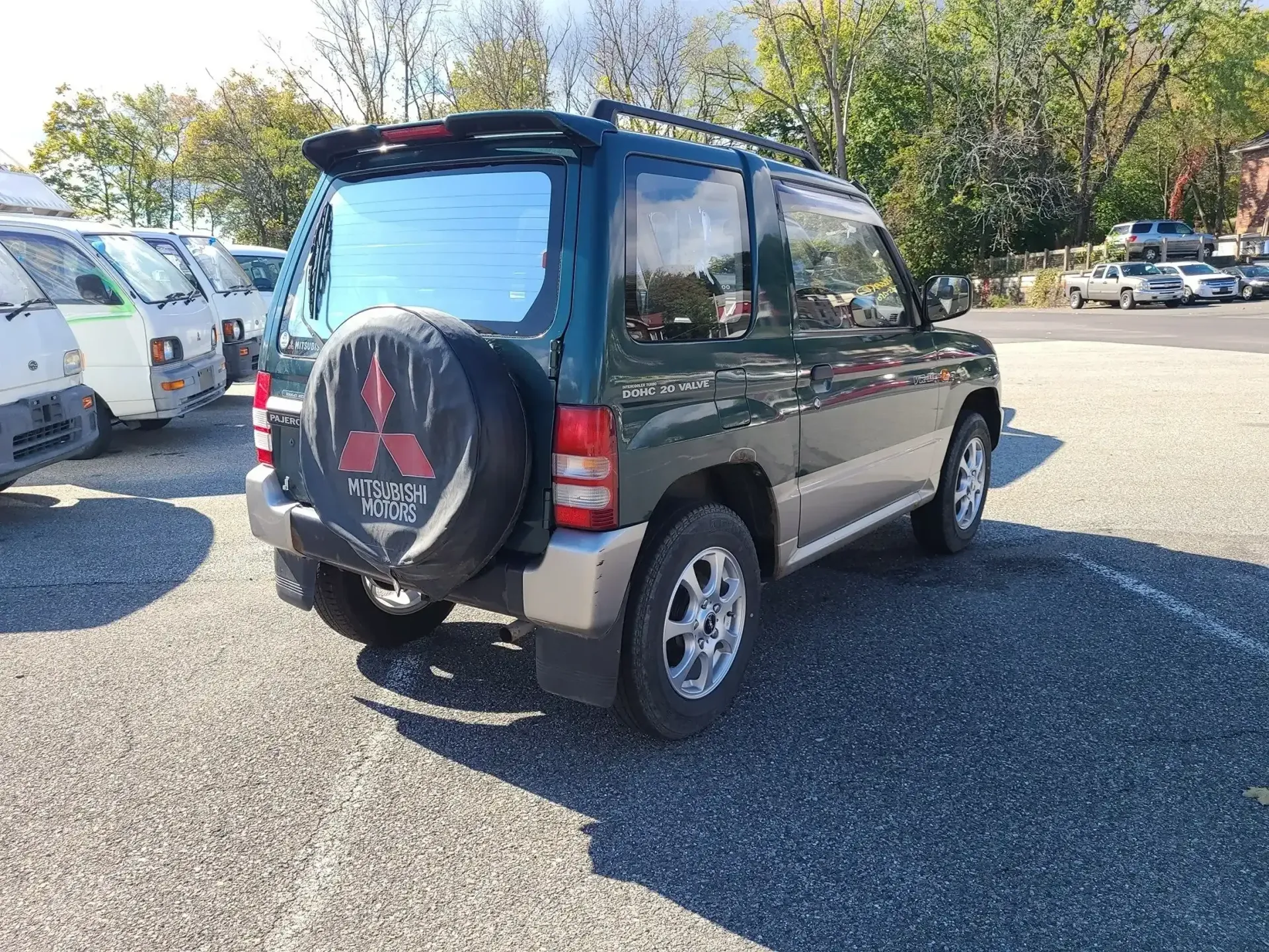 A dark green Mitsubishi Pajero Pinin with a spare tire cover, parked on a gravel lot under a bright, sunny sky.