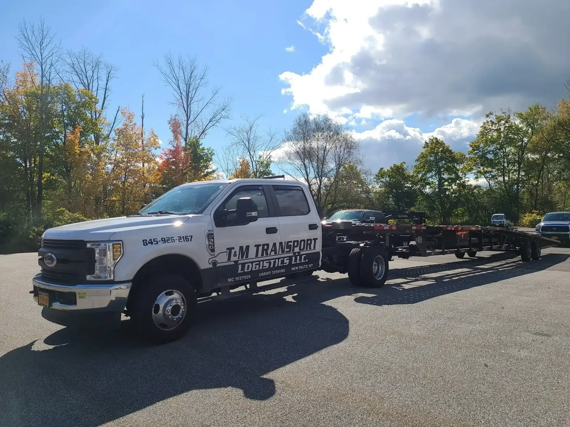 A white Ford pickup truck towing an empty flatbed trailer in a gravel lot with a treeline under a blue sky.