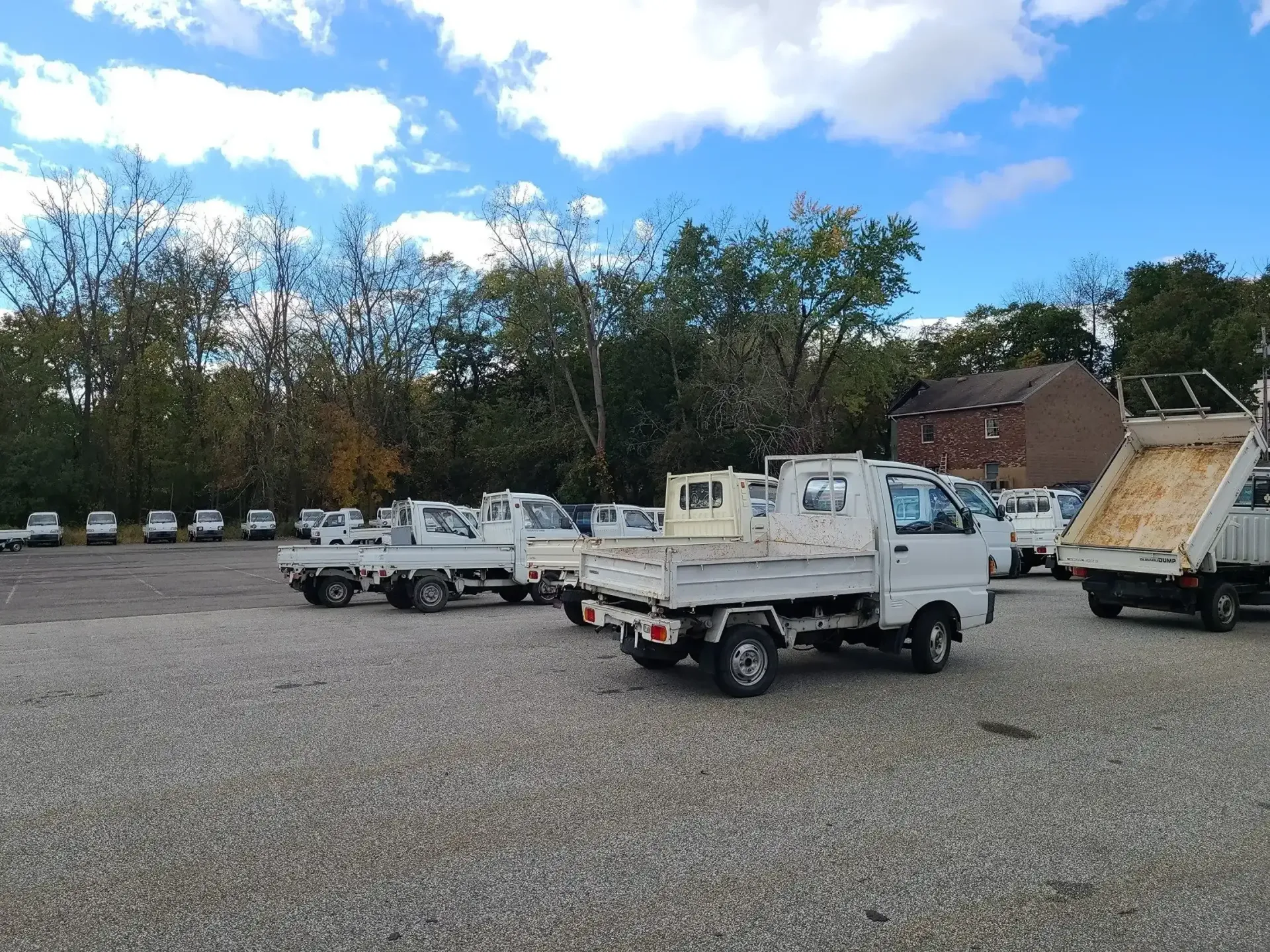 A row of white compact utility trucks parked on a gravel lot under a blue sky with scattered clouds.