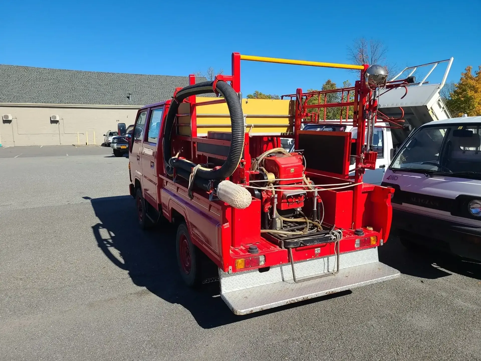 A bright red kei-style fire truck parked on a gravel lot, viewed from the rear, featuring hose equipment and a metal step.