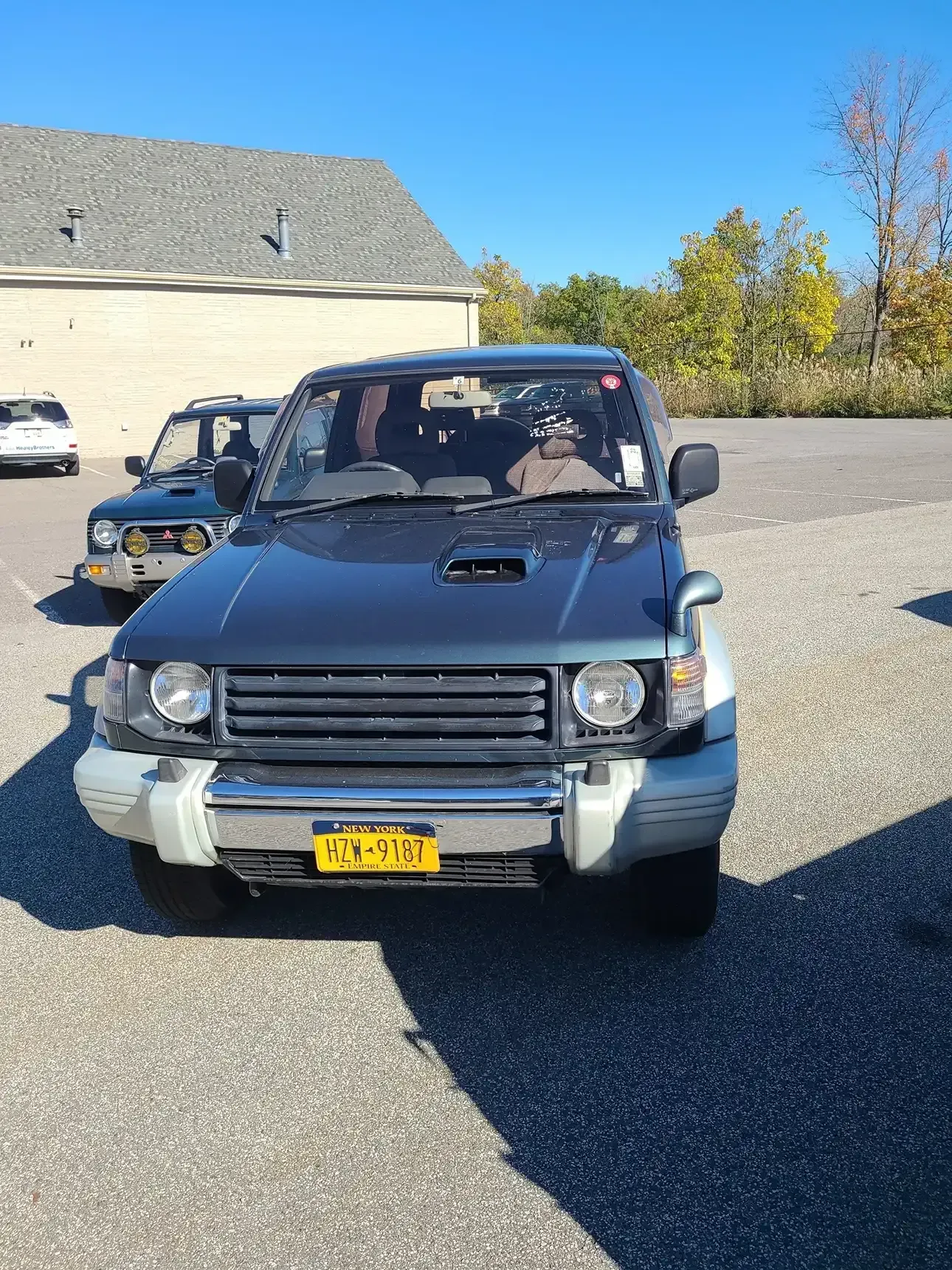 A dark green Mitsubishi Pajero SUV parked on a gravel lot under a clear blue sky, with a white vehicle behind it.