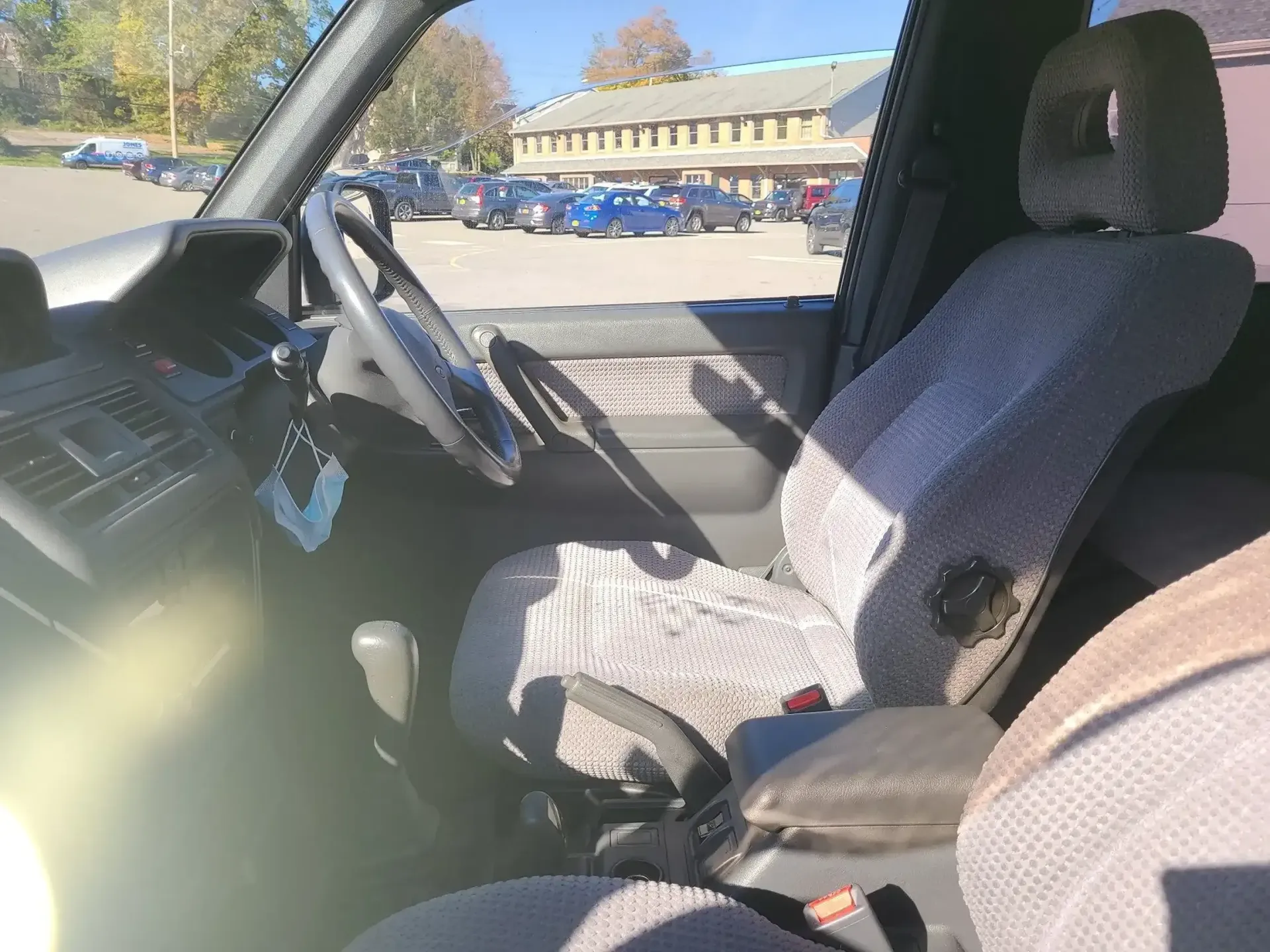 Interior view of a vehicle cab showing a textured gray driver's seat, steering wheel, and dashboard in a parking lot.