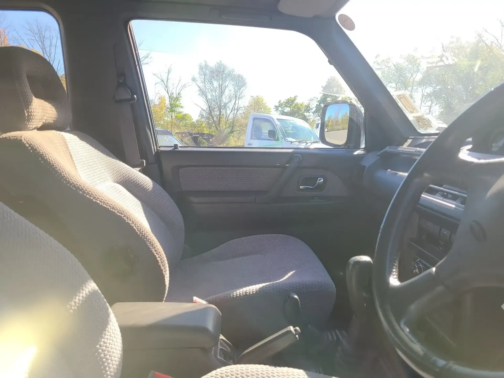 Interior view from the driver's side of a vehicle, showing grey fabric seats, a steering wheel, and an open side window.