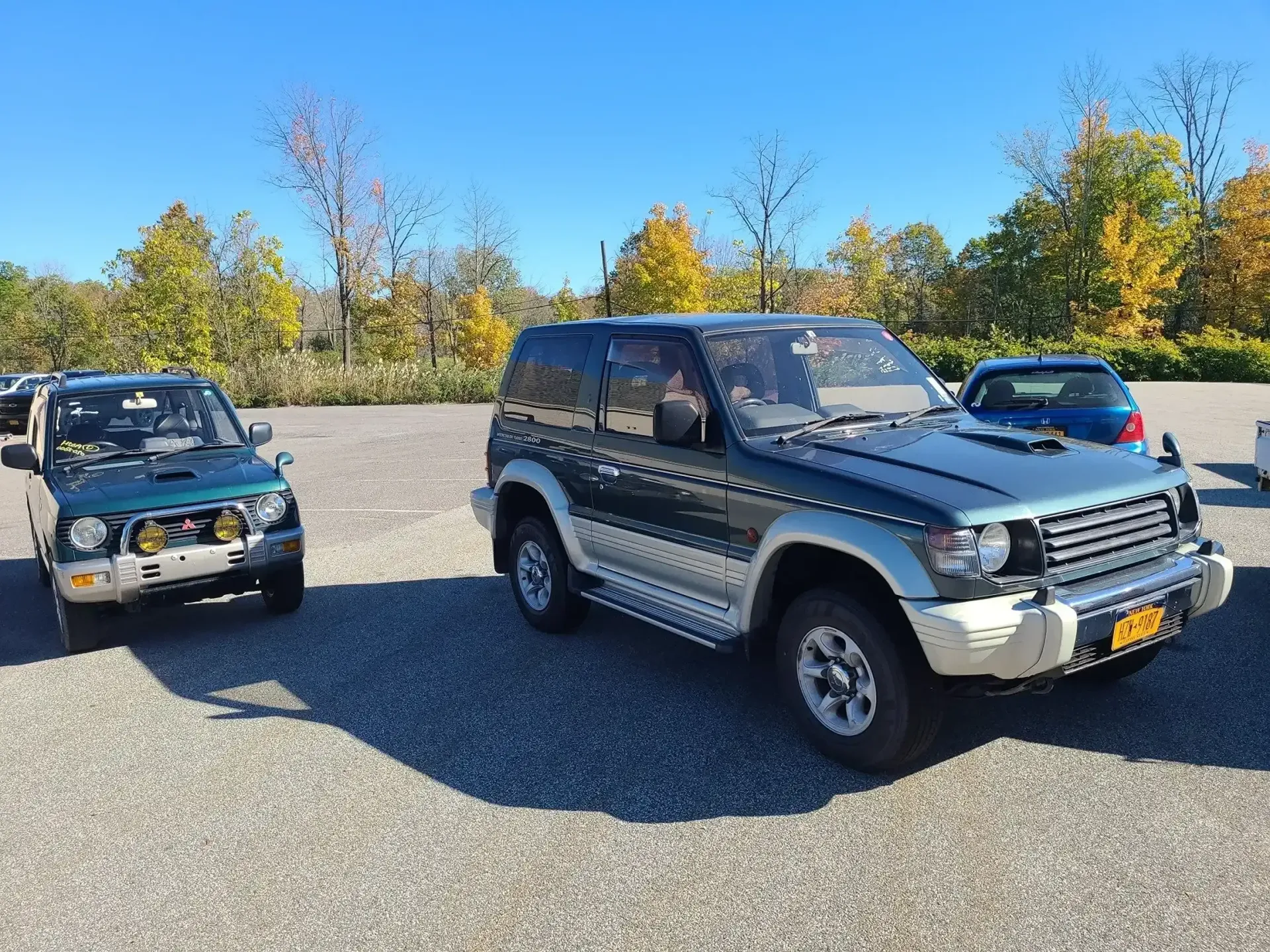 Two Mitsubishi Pajero SUVs parked on a gravel lot under a clear blue sky with autumn trees in the background.