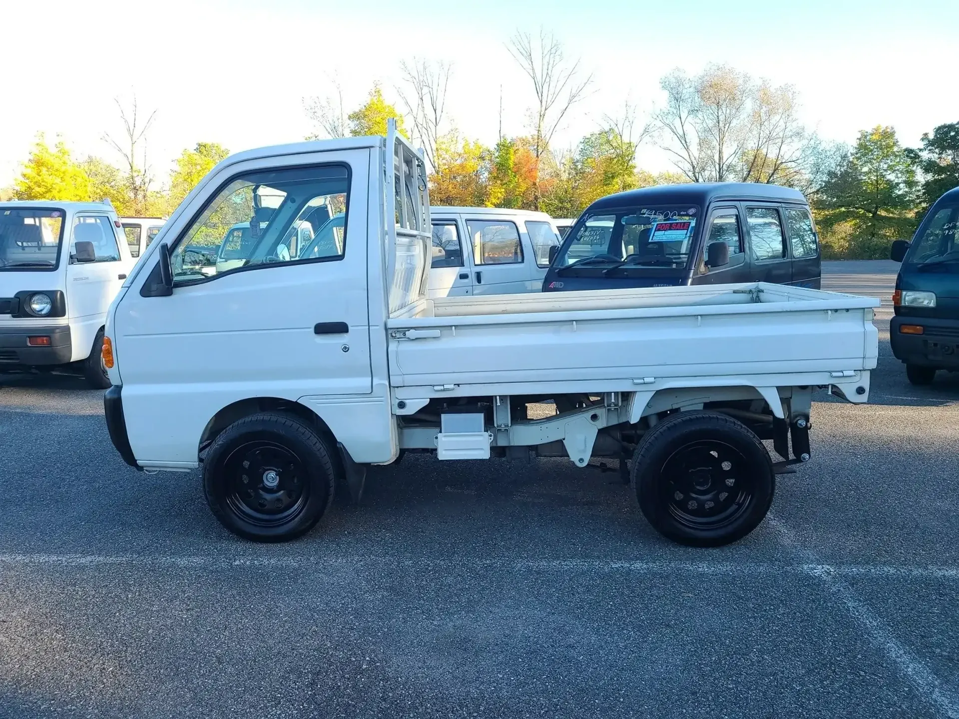A white Kei truck with black wheels parked in an outdoor gravel lot with other vehicles visible in the background.