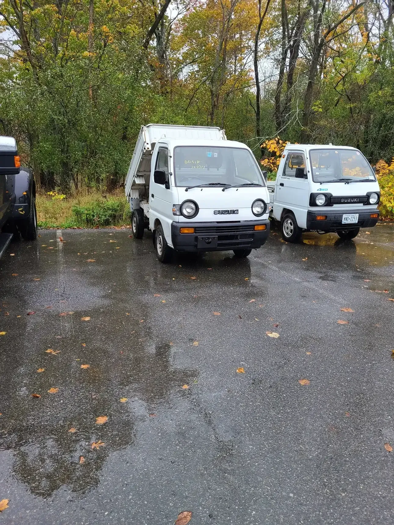 Two white small utility trucks parked side-by-side on a wet, paved lot with autumn trees in the background.