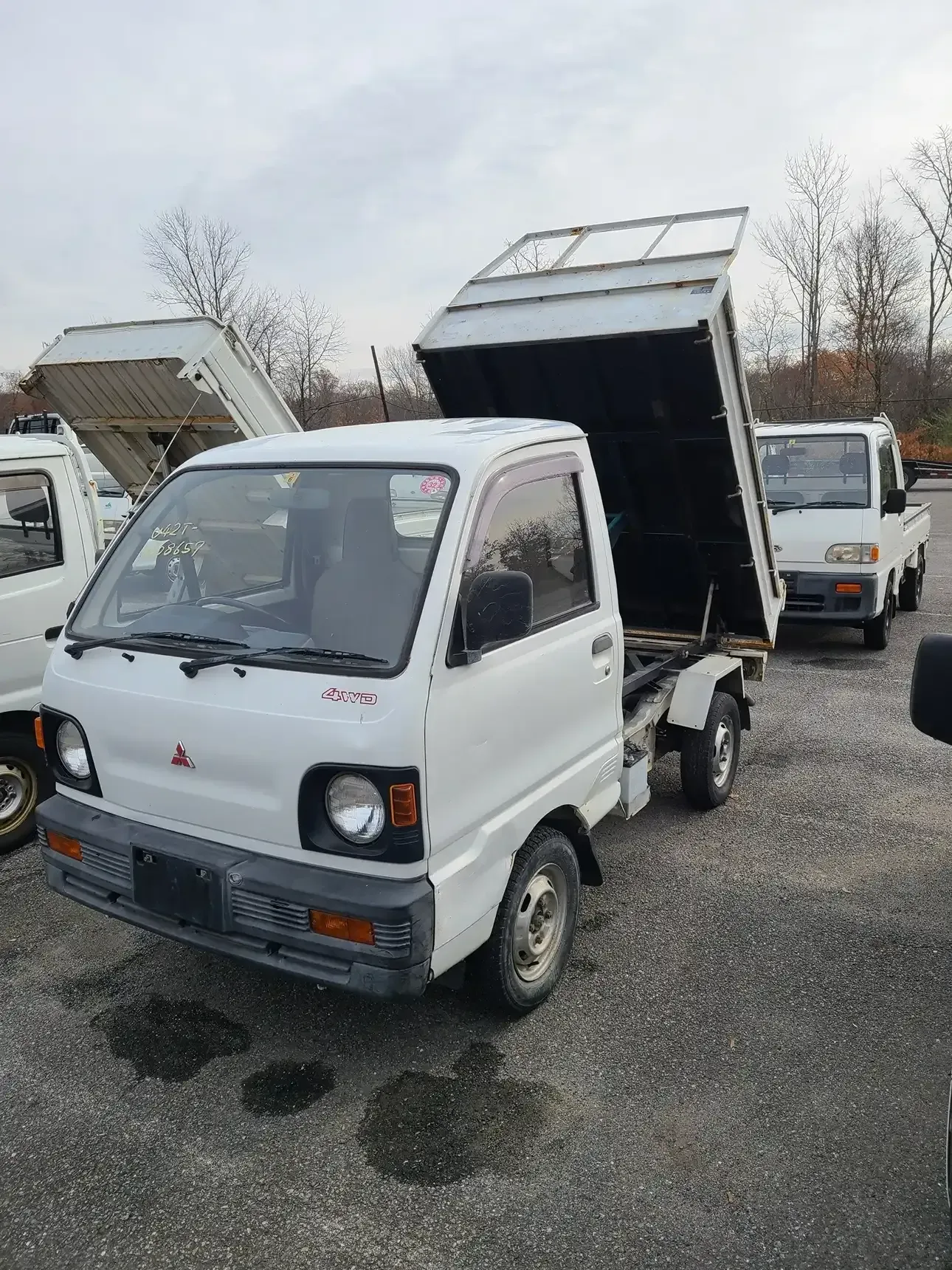 A white Mitsubishi mini dump truck with its bed raised, parked on a gravel lot with other similar trucks nearby.