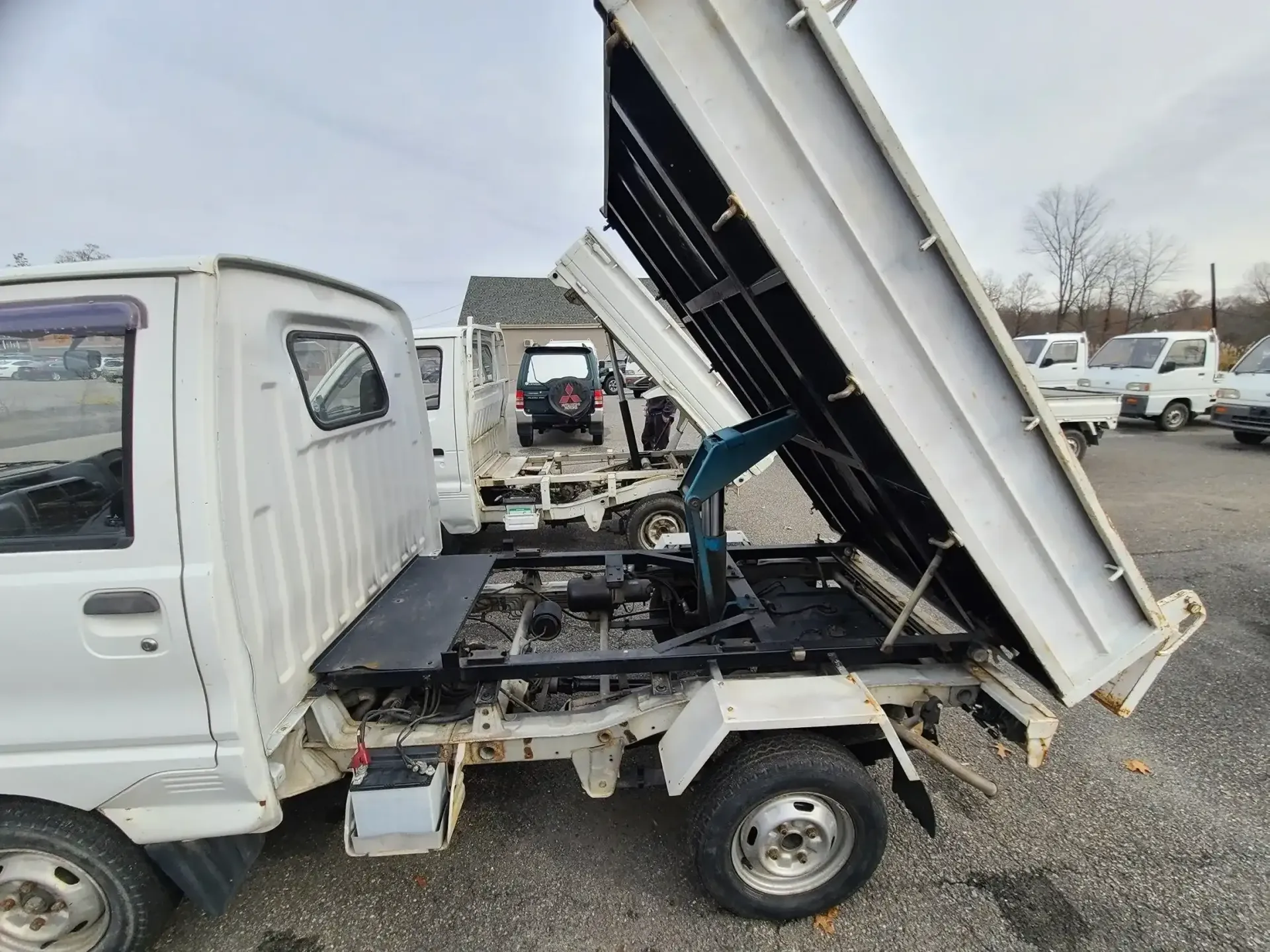 A white, small dump truck with its bed lifted up, parked in a lot with other similar vehicles.