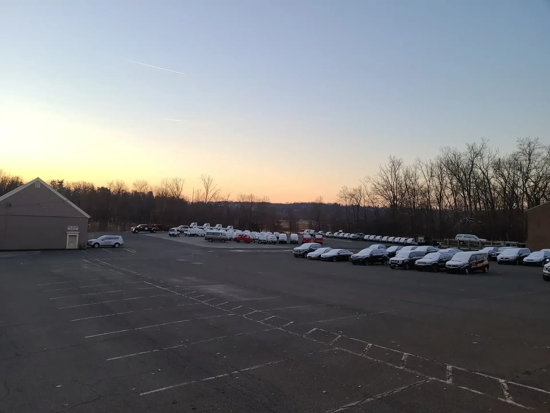 A parking lot filled with snow-covered cars during a sunset with a building on the left and trees in the background.