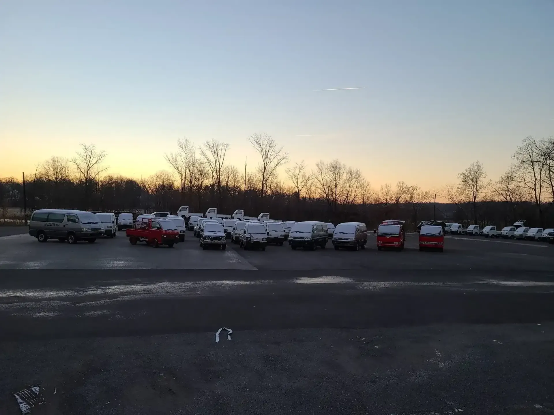 A parking lot filled with rows of vans and vehicles covered in light snow under a sunrise sky.