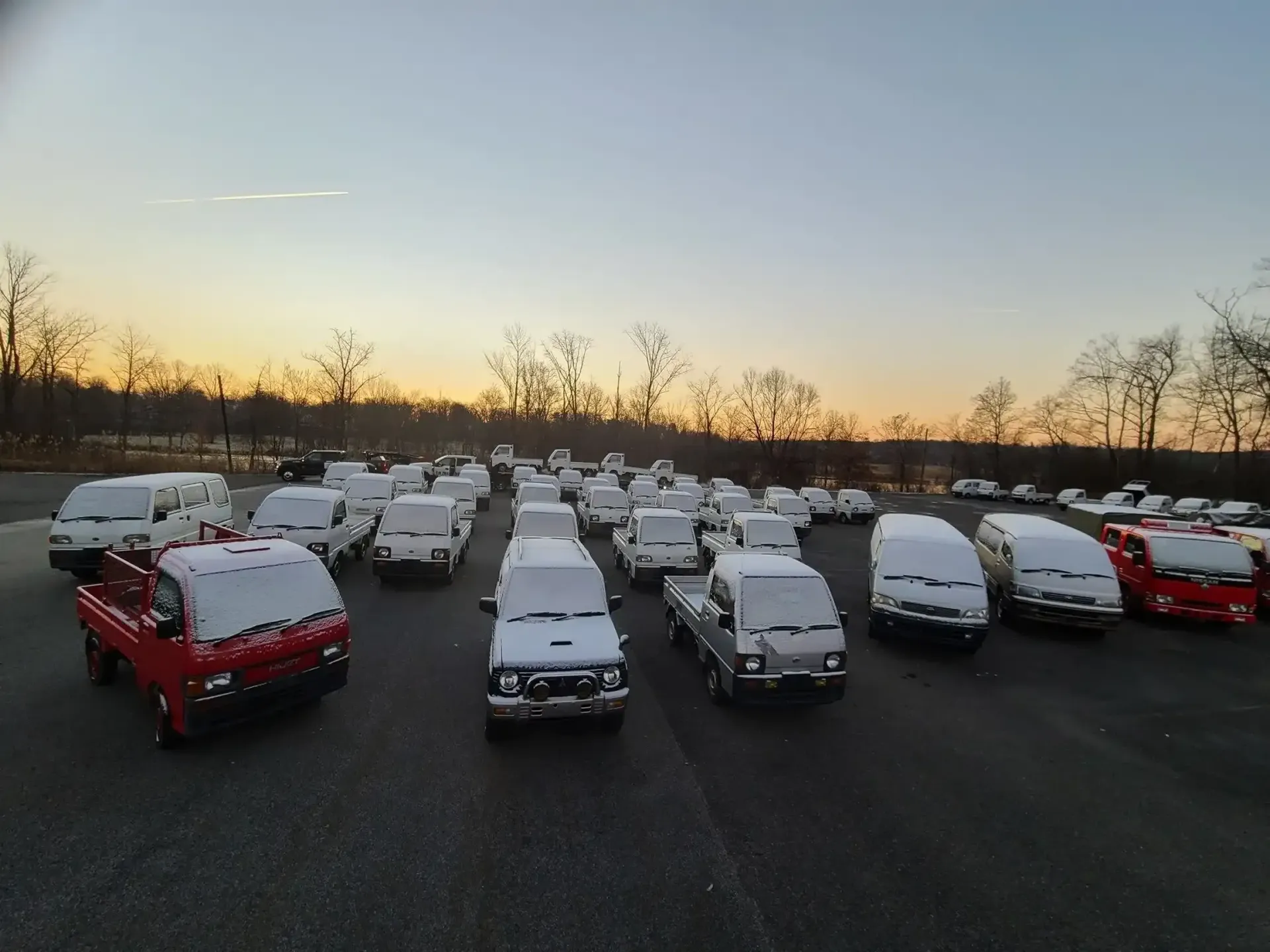 A large fleet of small, snow-covered trucks parked in a lot during sunset, with a tree line in the background.