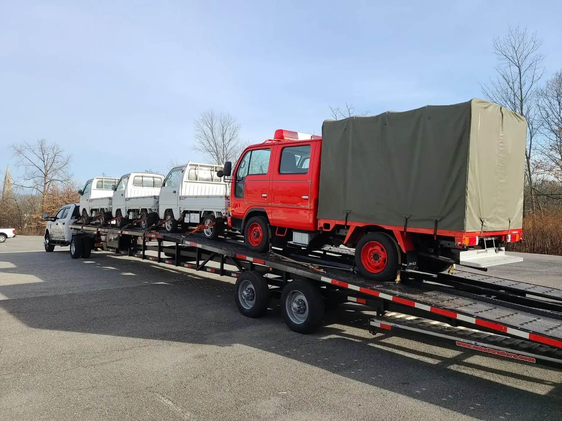 A red truck and three smaller vehicles loaded onto a flatbed trailer in a gravel lot on a sunny day.