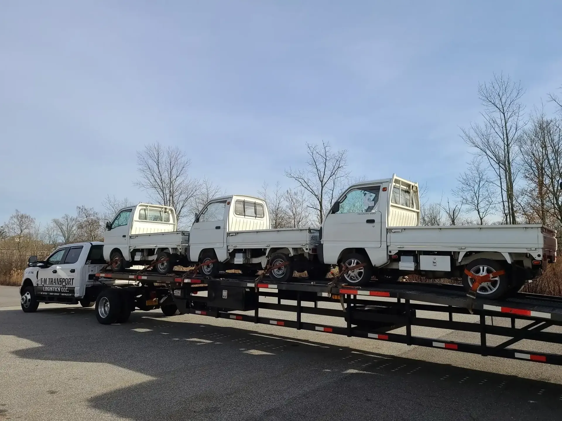 A flatbed trailer parked outdoors carrying three white Kei trucks against a clear blue sky.