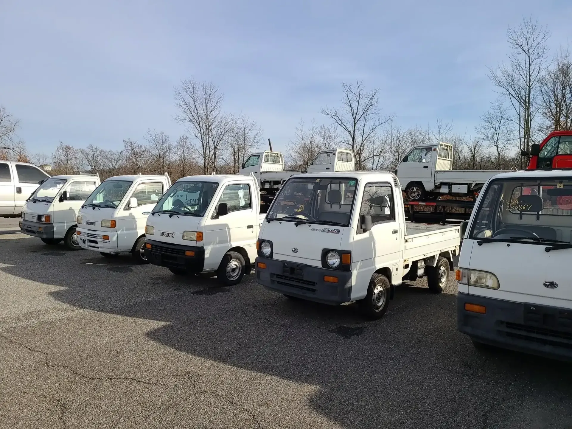 A row of white, small-sized flatbed trucks parked on a gravel lot under a bright sky.