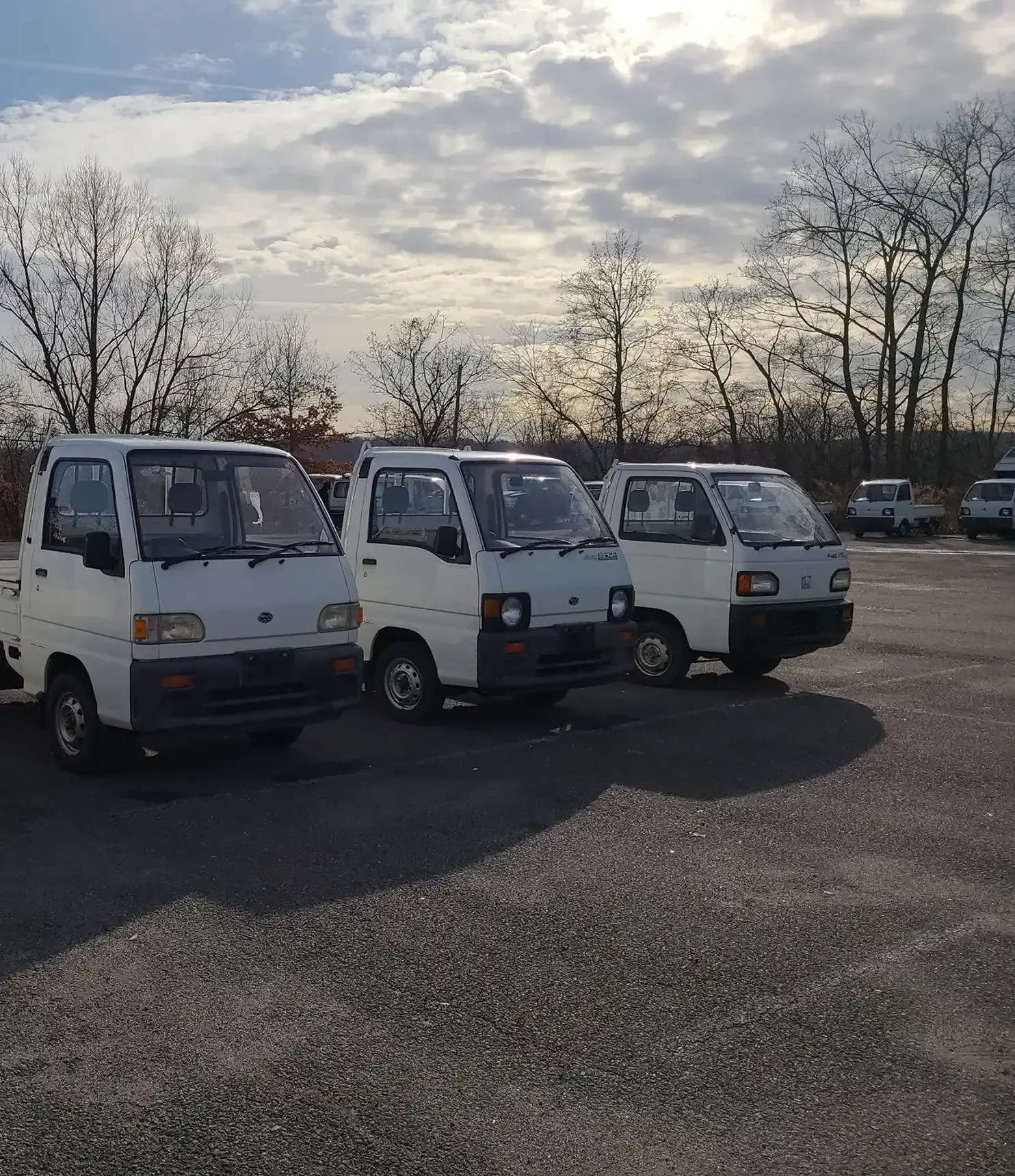 Three white Kei trucks parked in a gravel lot under a bright, cloudy sky.