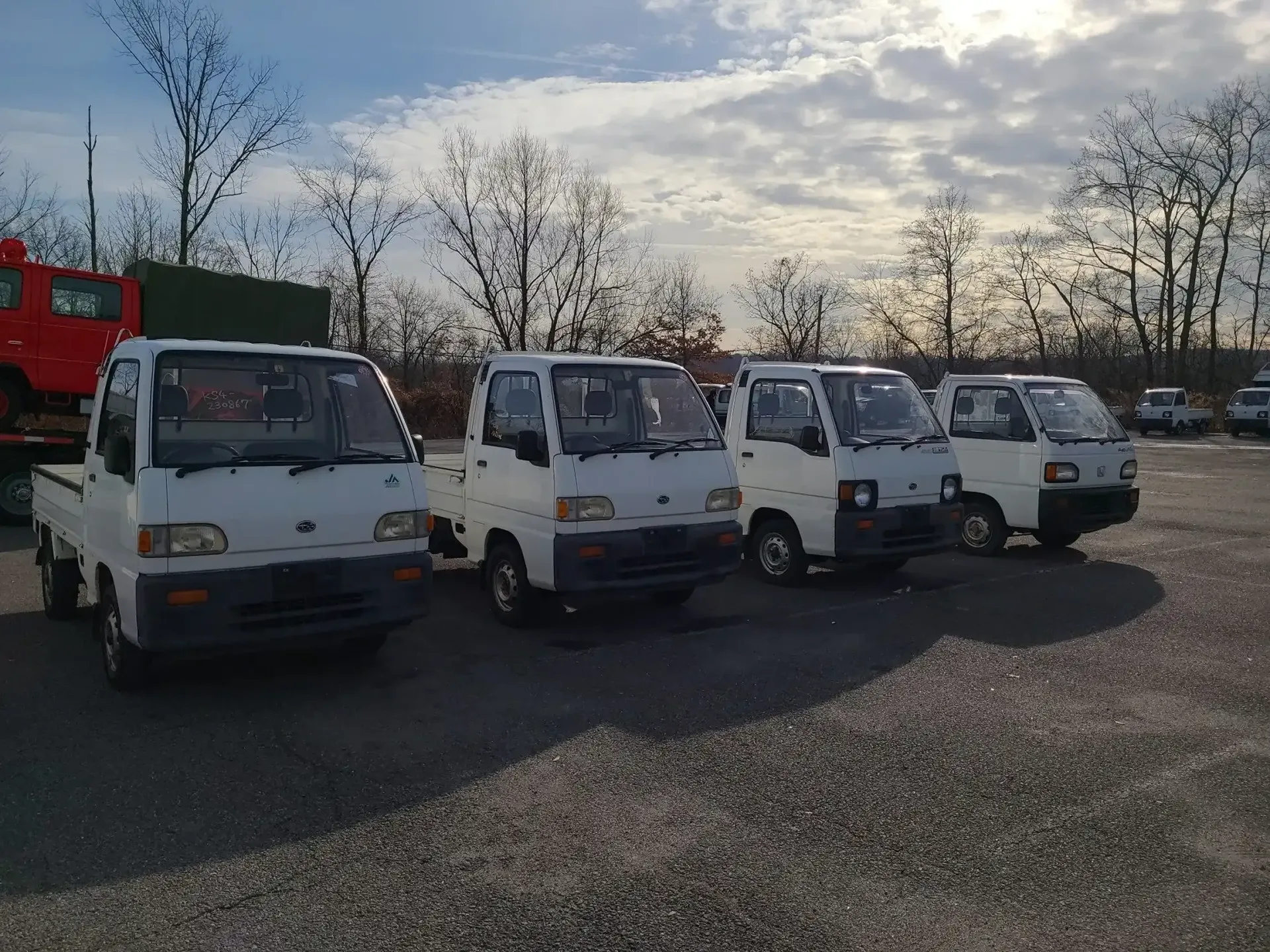 A row of four white mini trucks parked in an outdoor gravel lot under a bright, cloudy sky.