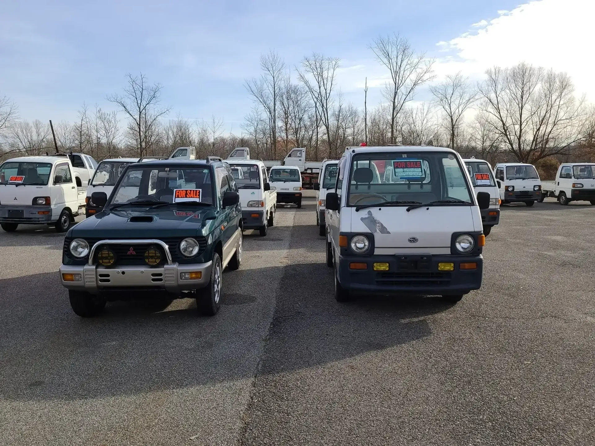 A fleet of small Japanese Kei trucks and a dark green SUV parked in a gravel lot under a bright, cloudy sky.