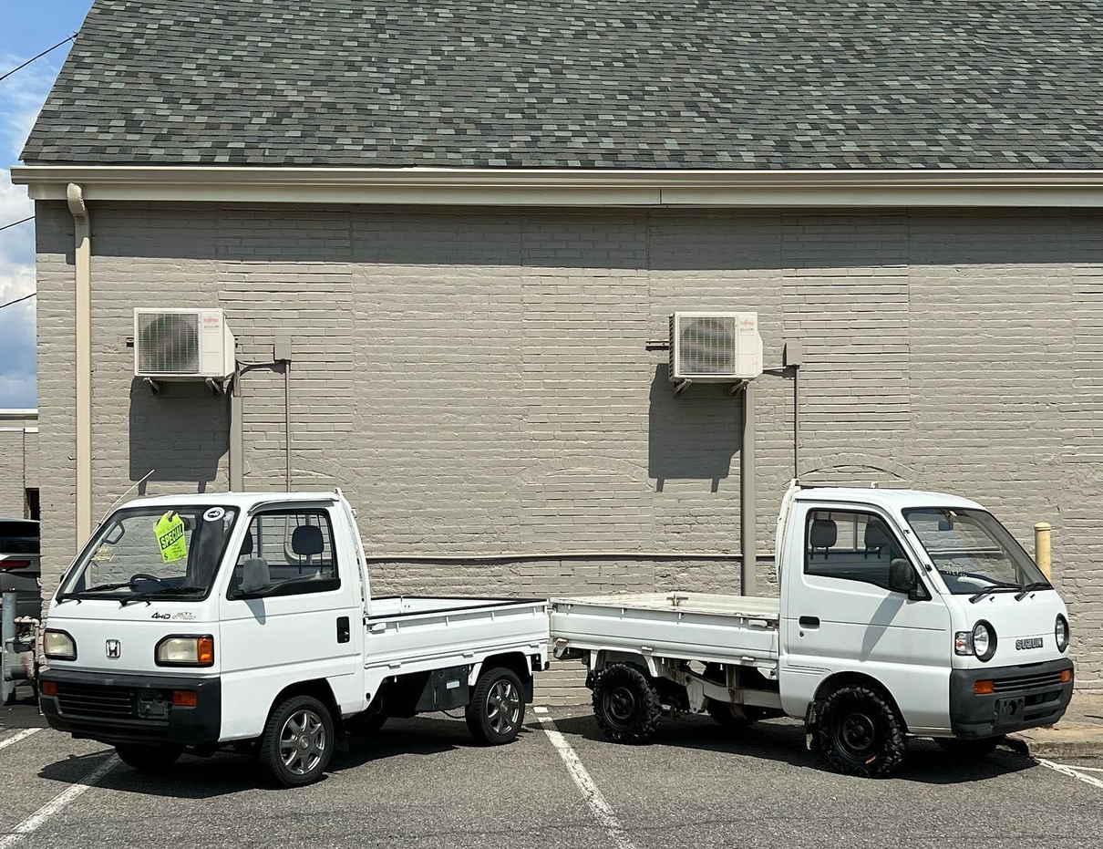 Two white mini-trucks parked side-by-side in front of a brick building with two air conditioning units.