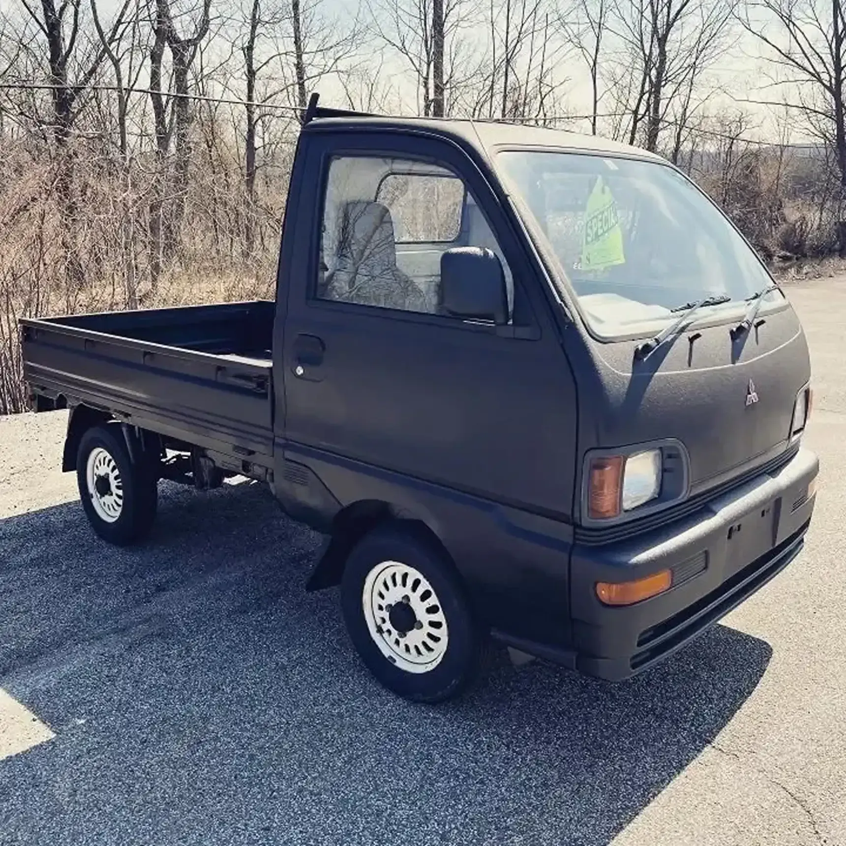 A matte black Mitsubishi kei truck parked on an asphalt lot with bare trees in the background.