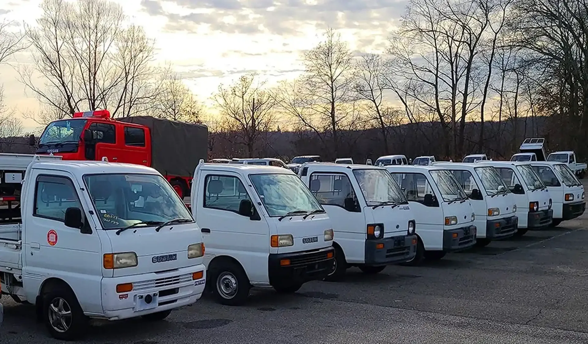 A line of white mini-trucks parked in an outdoor lot at dusk, with a red fire truck visible in the background.
