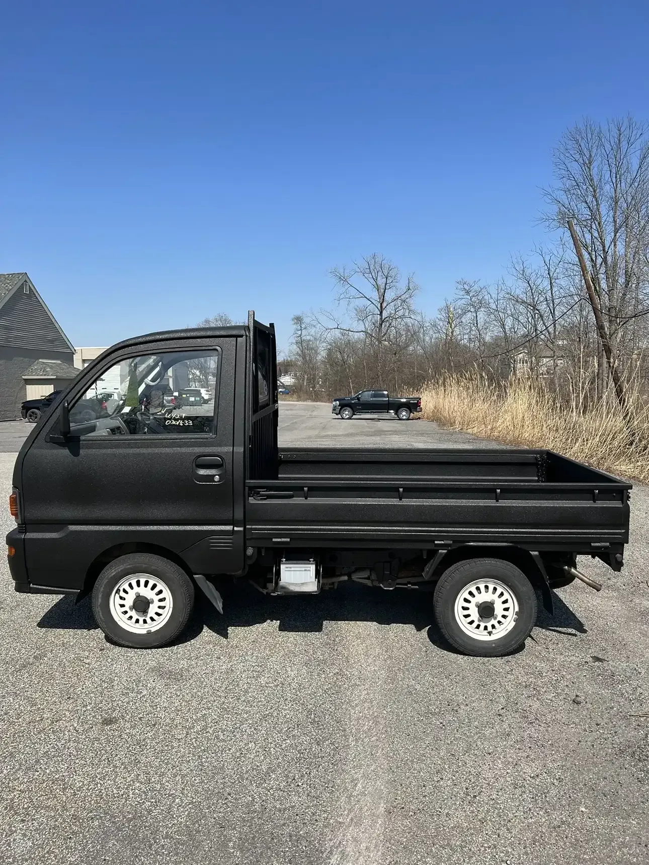 A side view of a small black pickup truck with white wheels parked on a gravel lot under a clear blue sky.