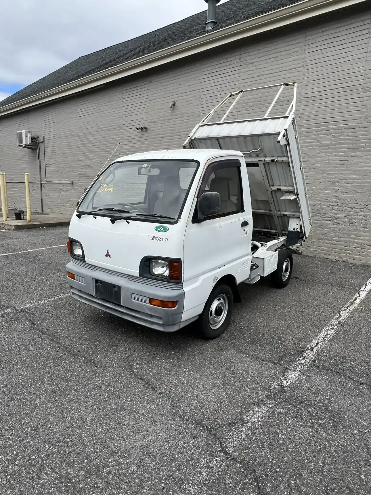 A white Mitsubishi kei dump truck parked in an asphalt parking lot next to a light-colored brick building.