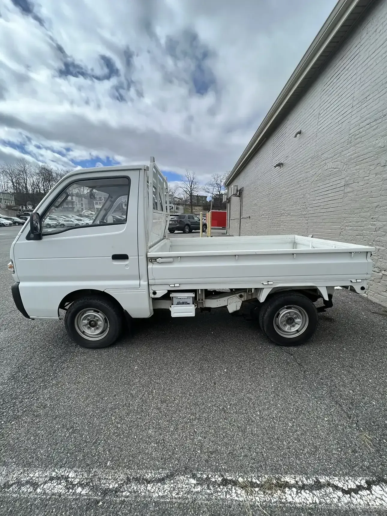 A white Kei mini truck parked on an asphalt lot beside a white brick building under a cloudy sky.