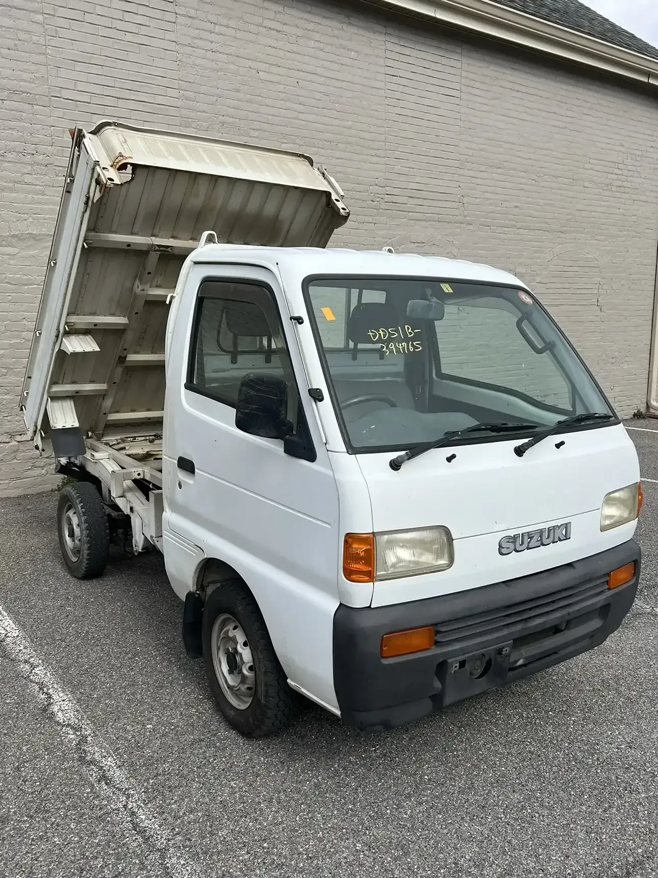 A white Suzuki Carry dump truck parked on asphalt in front of a light-colored brick wall.