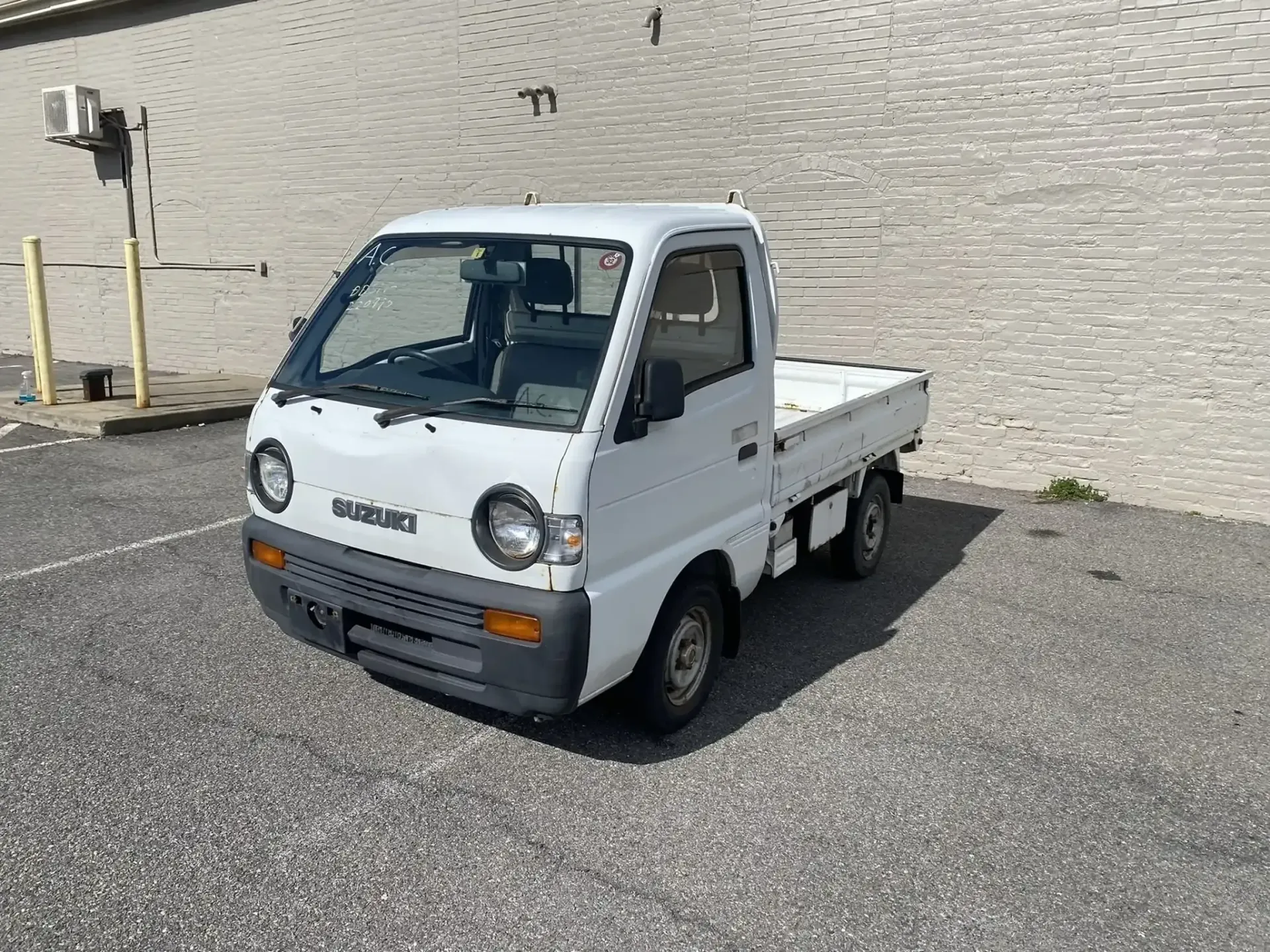 A small white kei truck parked on an asphalt lot in front of a beige masonry wall.