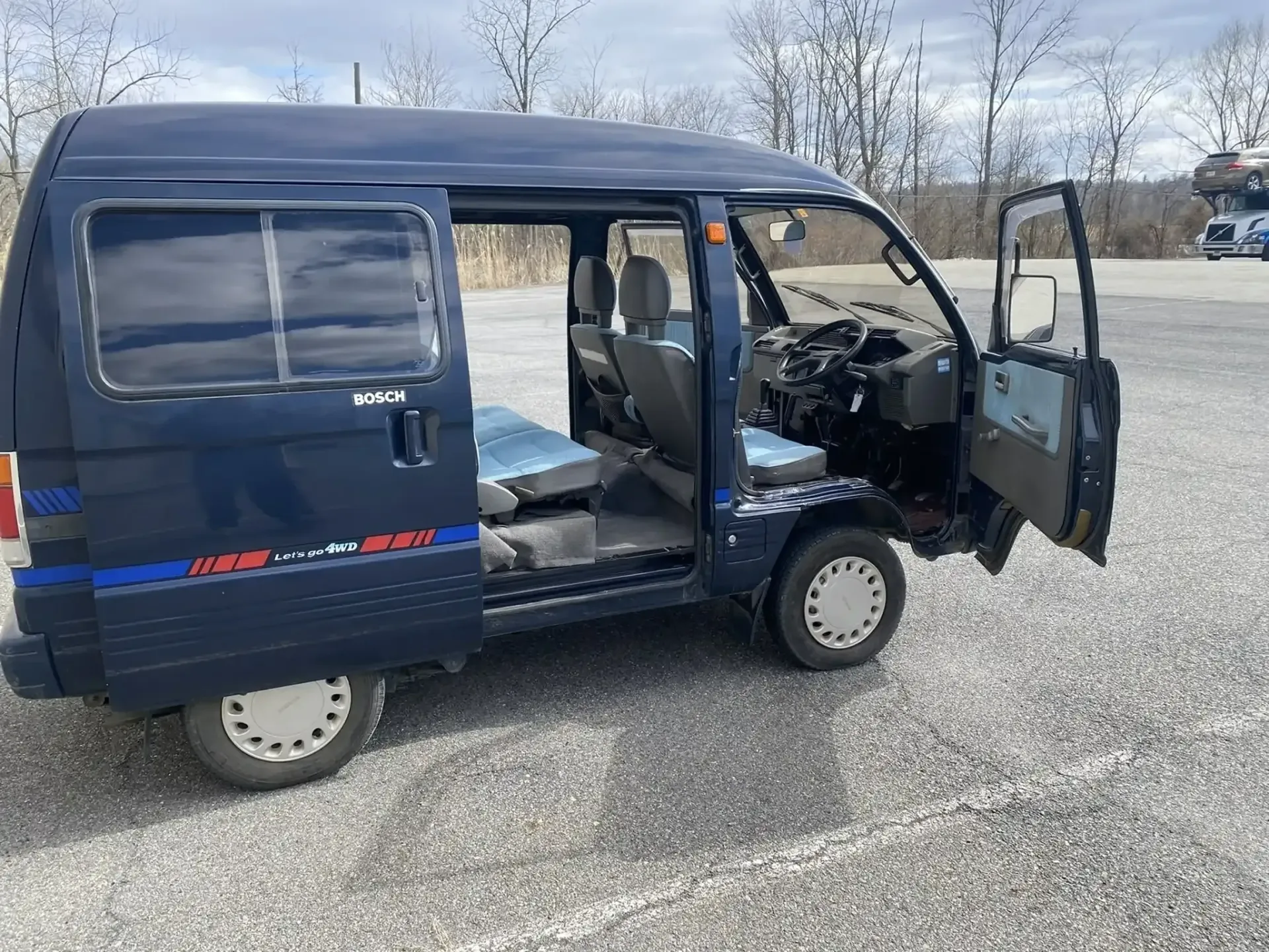 A blue Japanese kei van with an open side door and driver door, parked on a gravel lot under a cloudy sky.