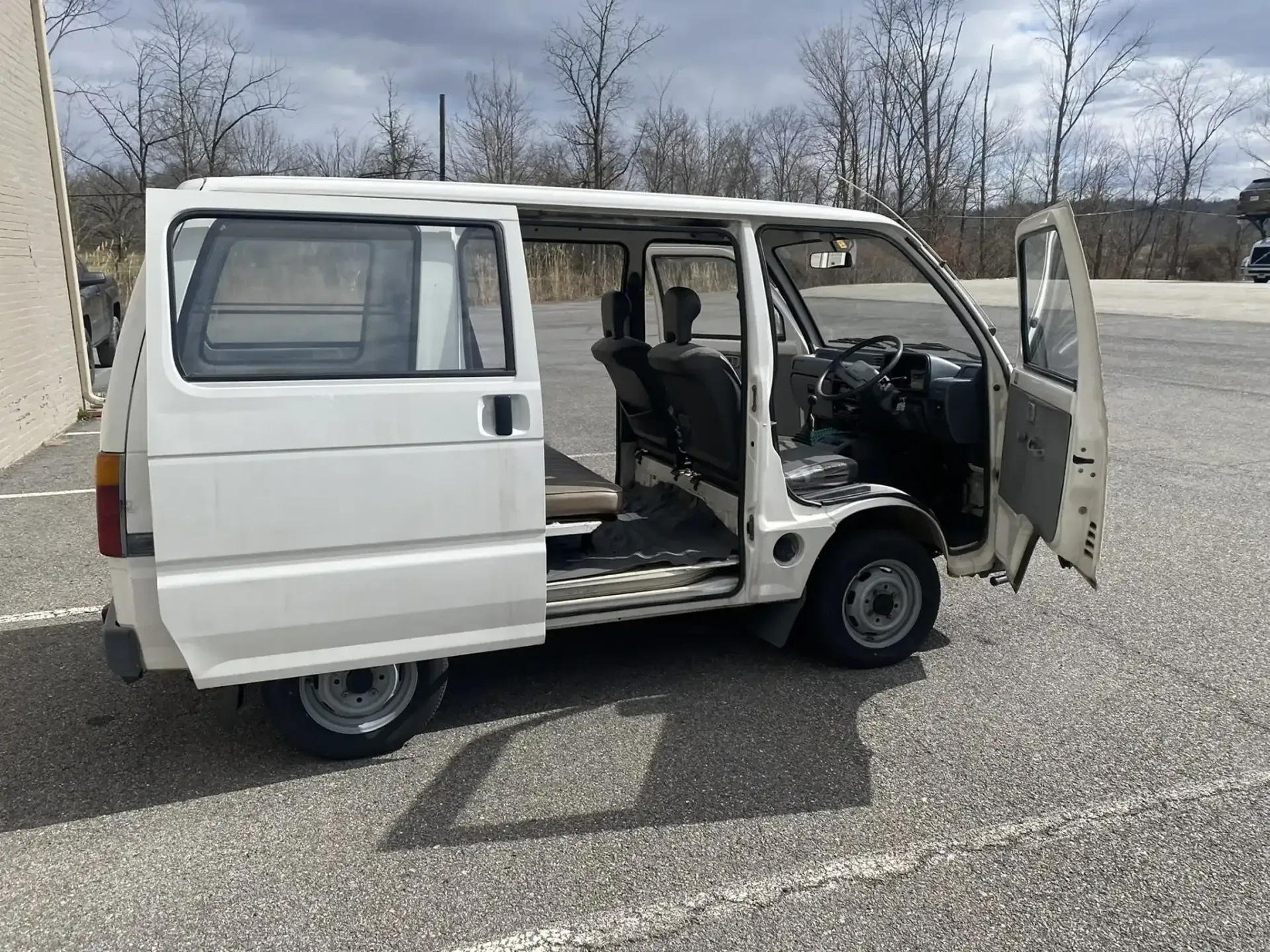 A white, compact Japanese kei van parked in an asphalt lot with its side sliding door and front door open.