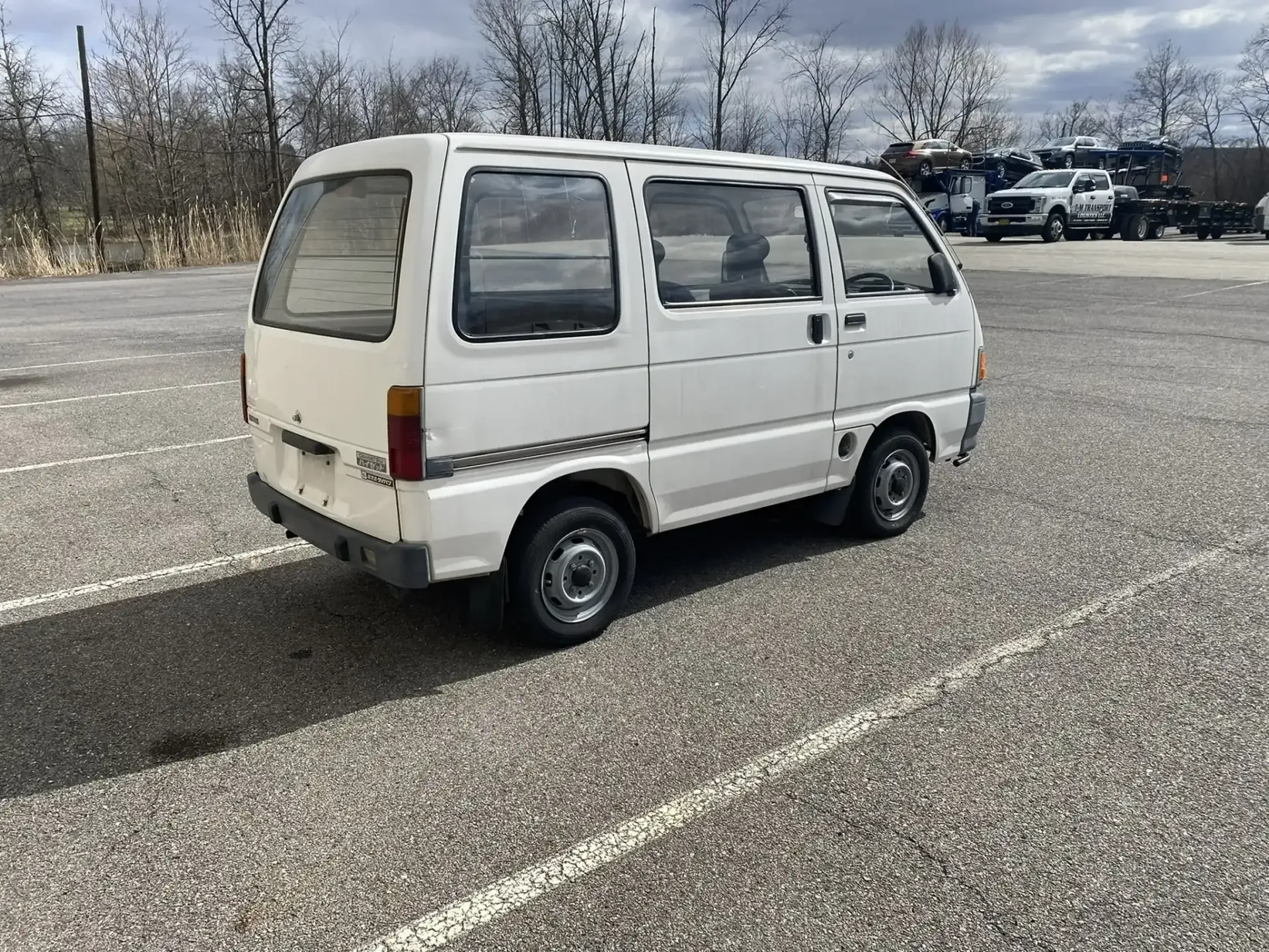 A white, boxy Kei van parked on a gravel lot under a cloudy sky.