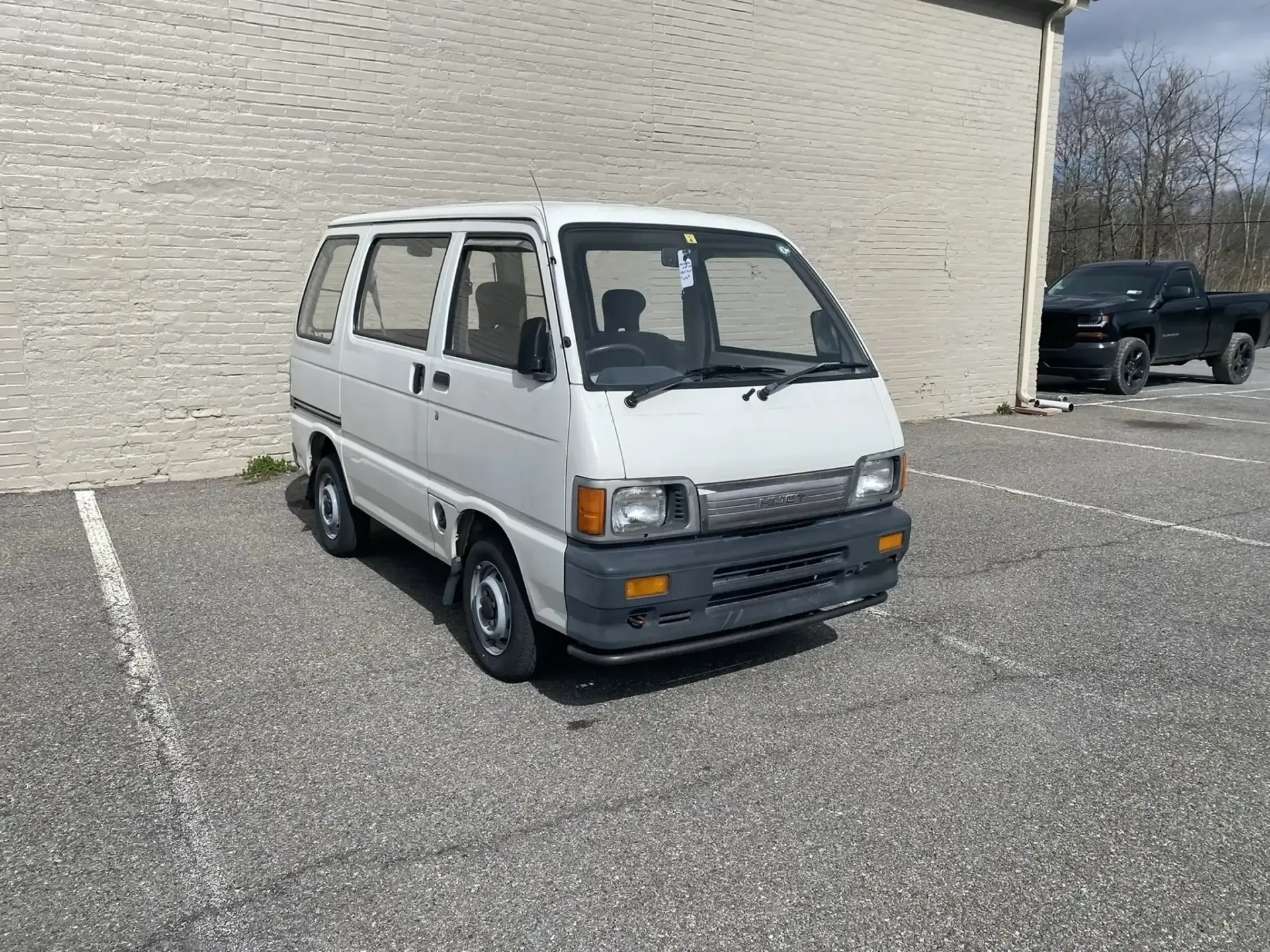 A white kei van parked in a gravel lot next to a brick building, with a dark pickup truck visible in the background.