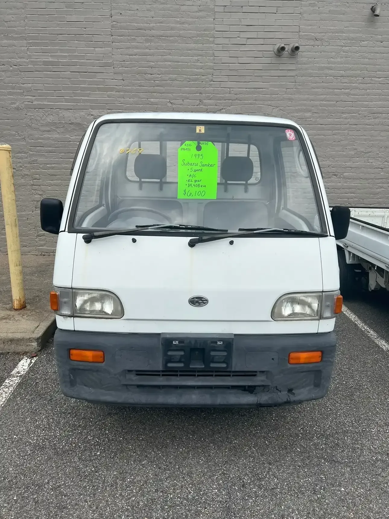 A white Subaru Sambar kei truck parked outdoors with a bright green information sign displayed in the windshield.