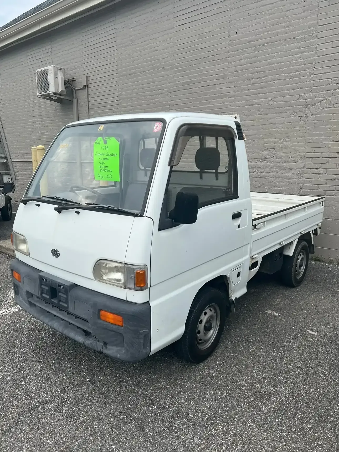 A white kei truck parked on asphalt in front of a gray brick building with a green sign in the windshield.