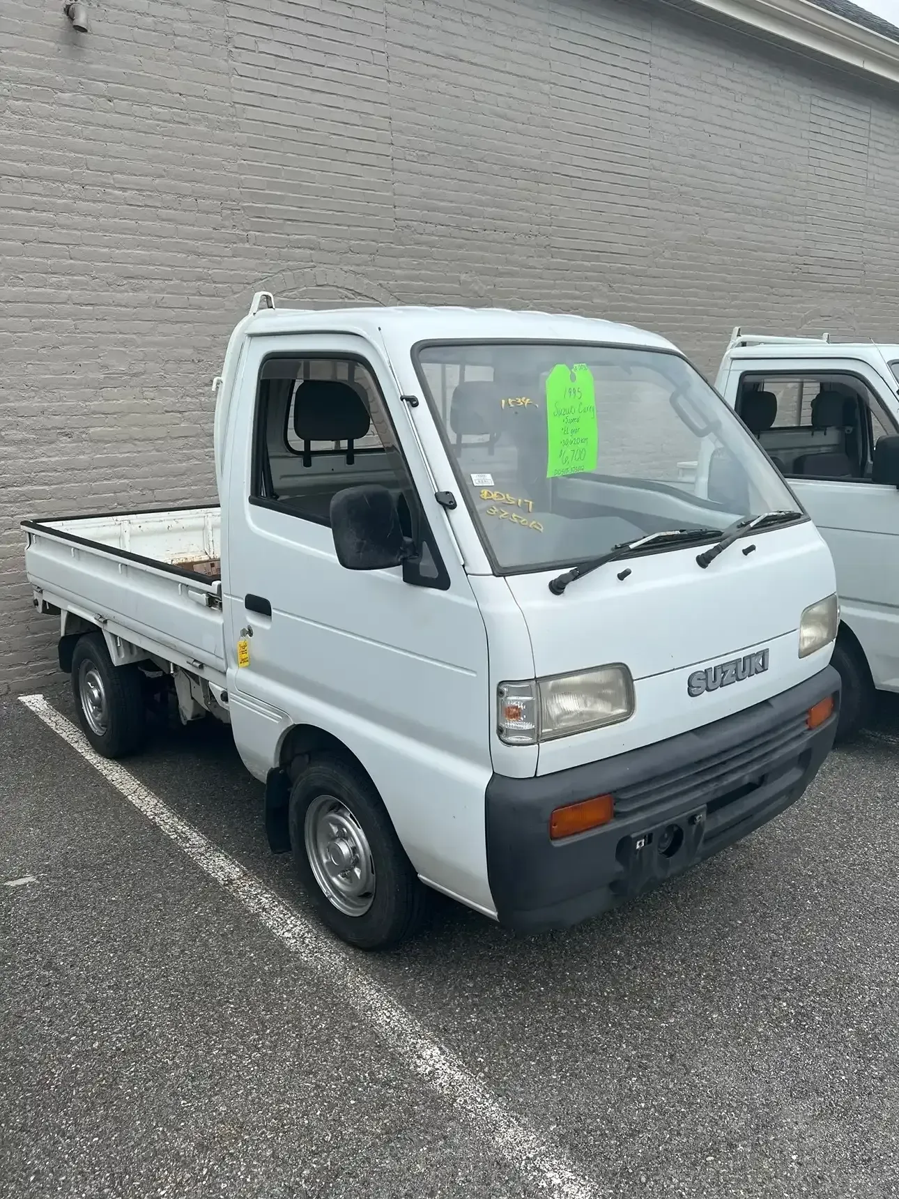 A white Suzuki mini-truck parked in an outdoor lot with a green sticker visible on the windshield.