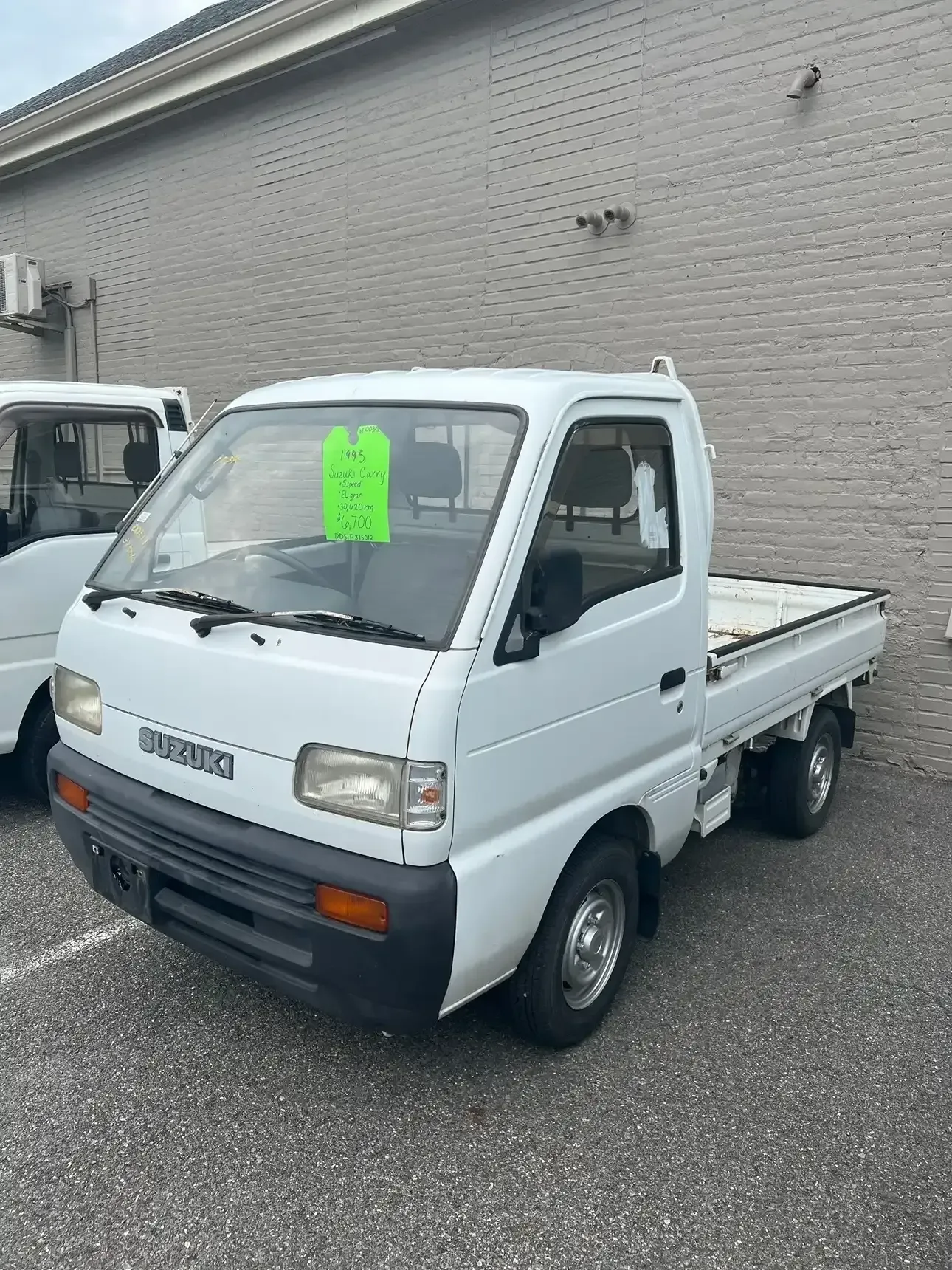 A white Daihatsu Hijet kei truck parked on a gravel lot next to a brick building.