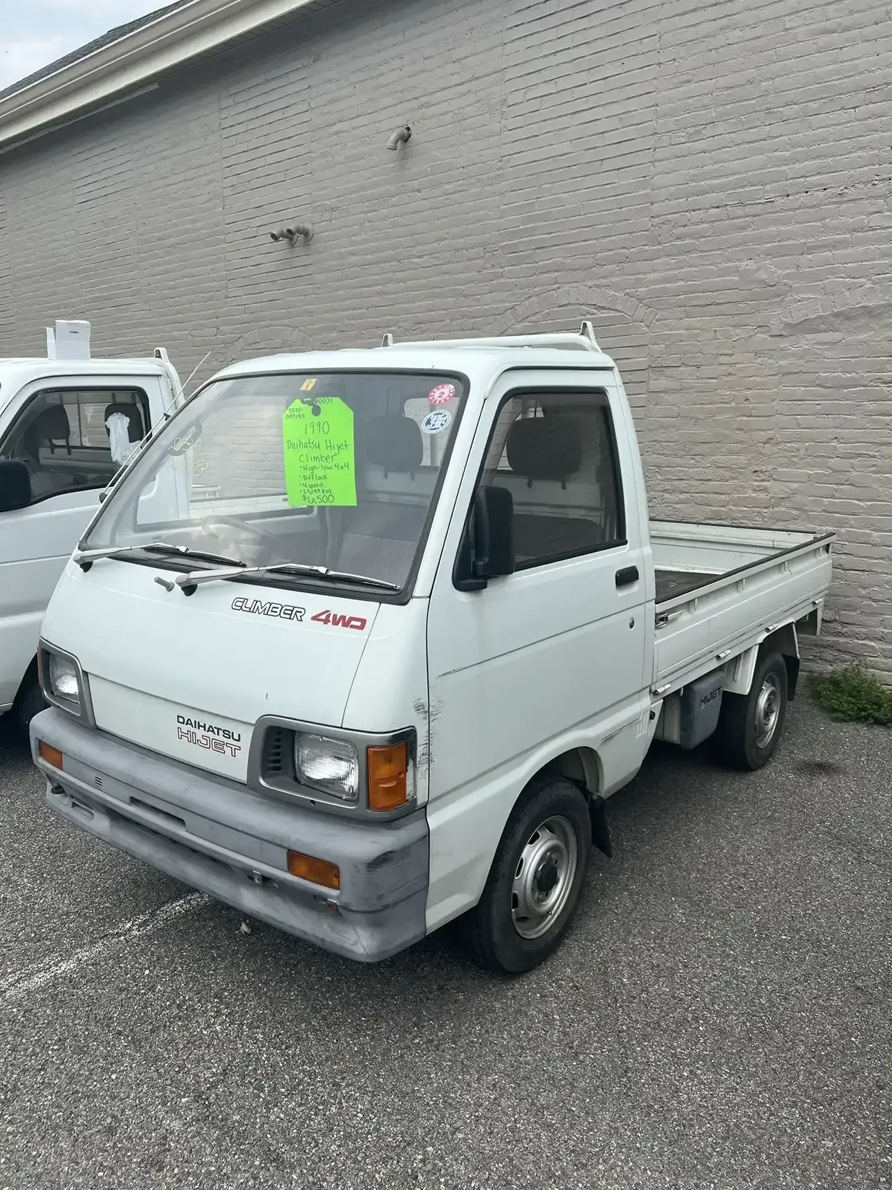 A white, small flatbed pickup truck parked on an asphalt lot in front of a brick building.