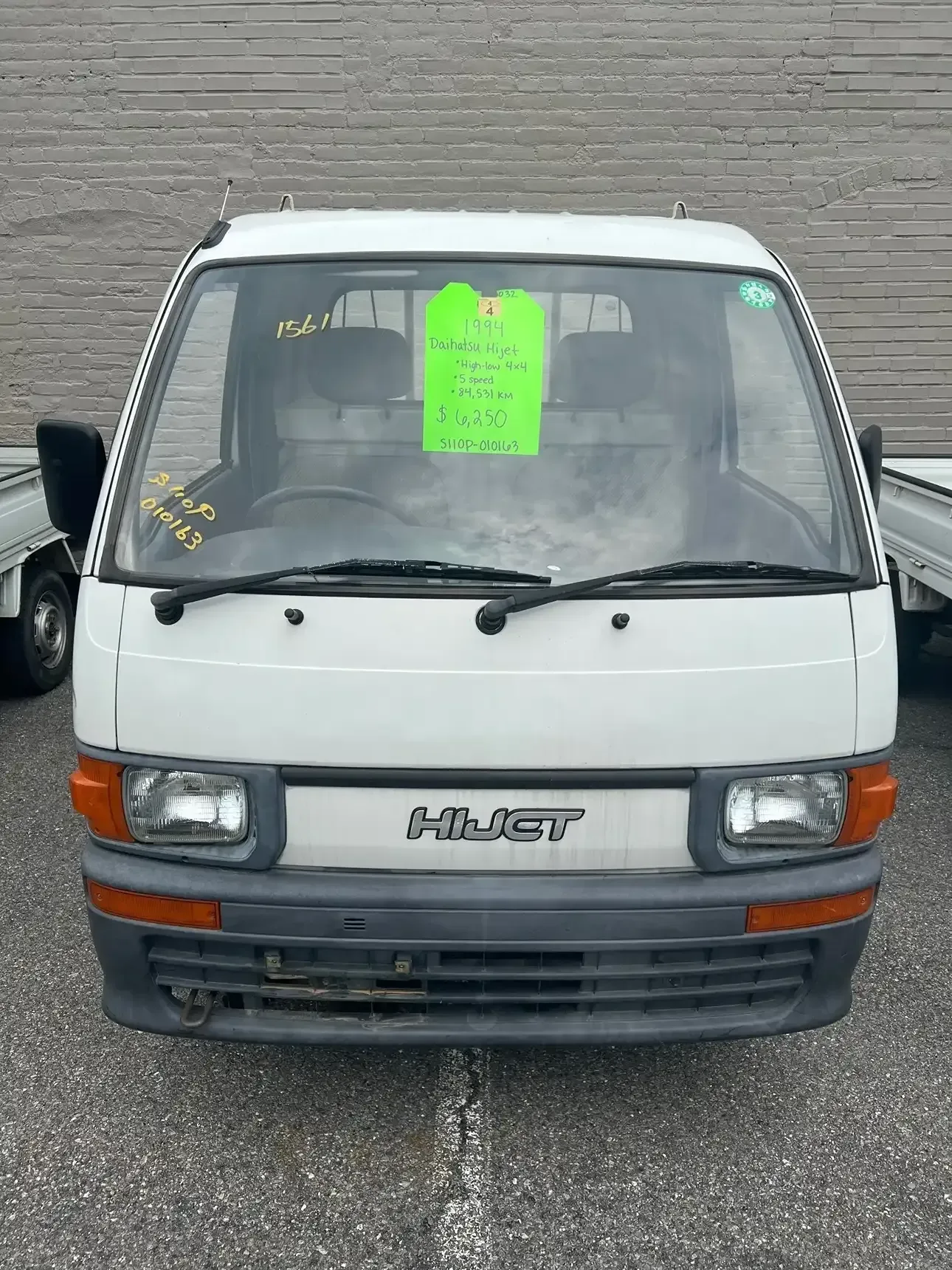 Front view of a white Daihatsu Hijet kei truck with a green sales sign on the windshield, parked outdoors.