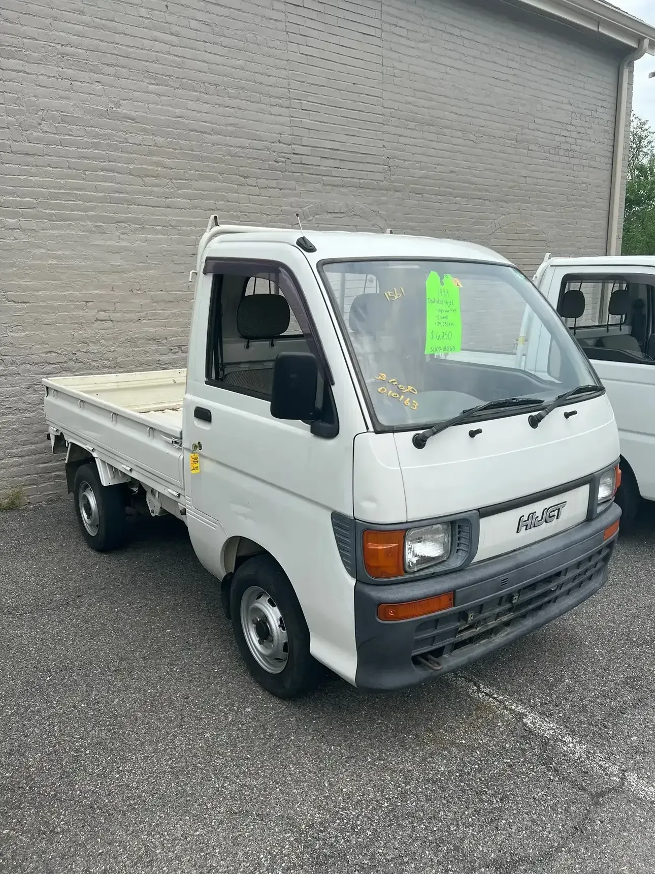 A white Kei truck parked on asphalt next to a building, with a green price tag on its windshield.