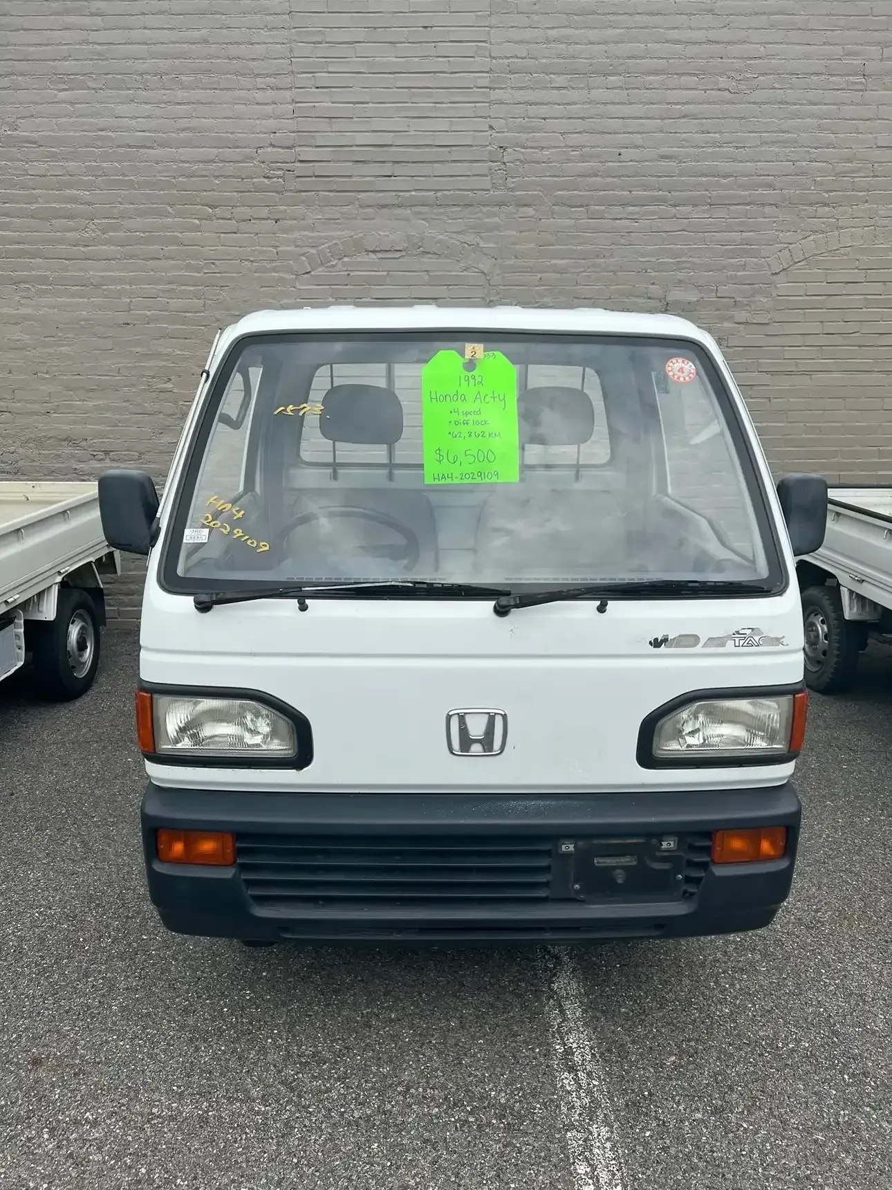 A white Honda Acty mini truck parked outdoors with a neon green sales tag attached to the interior windshield.