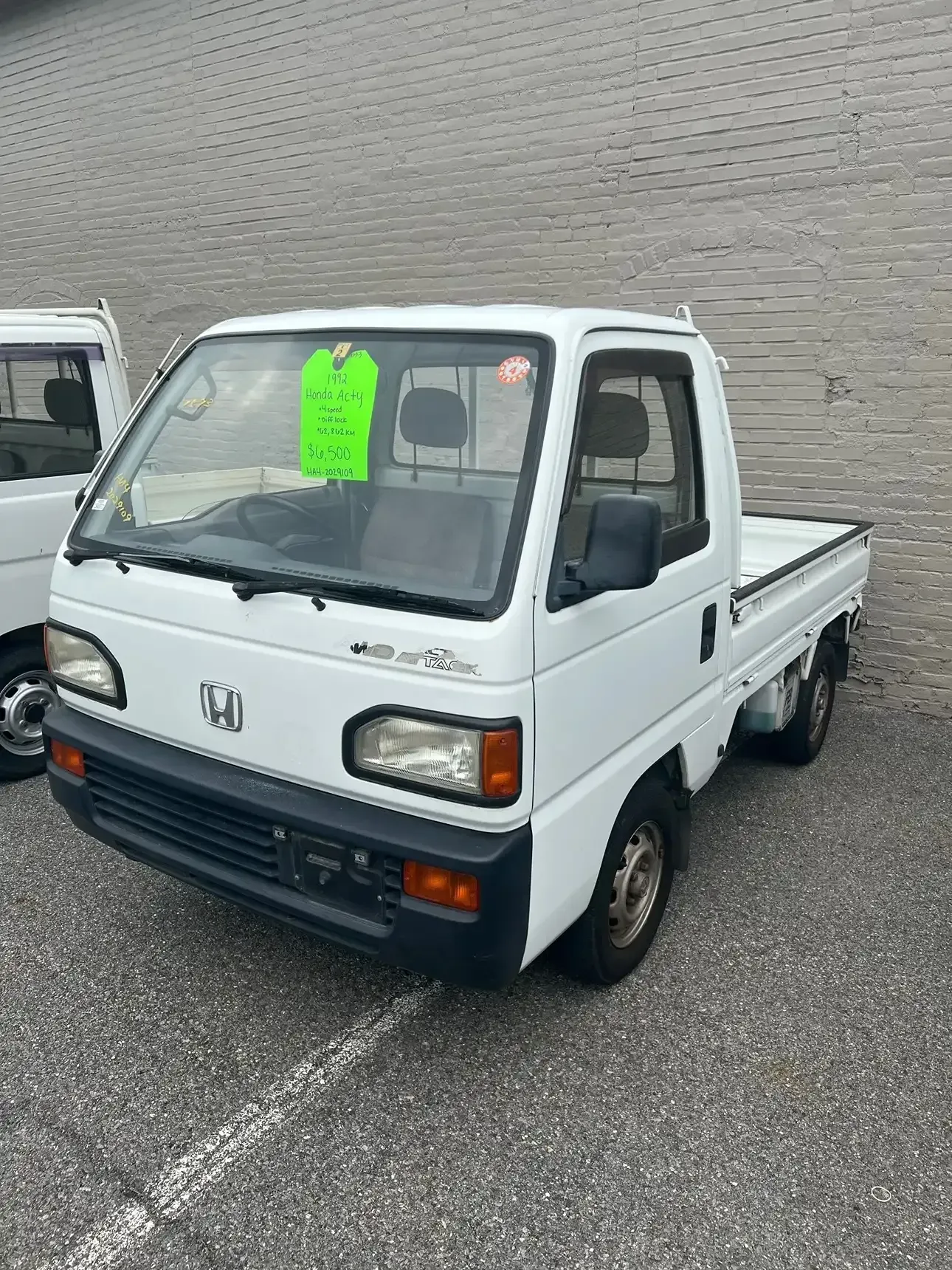 A white Honda Acty kei truck parked on an asphalt lot with a green price tag on the windshield.