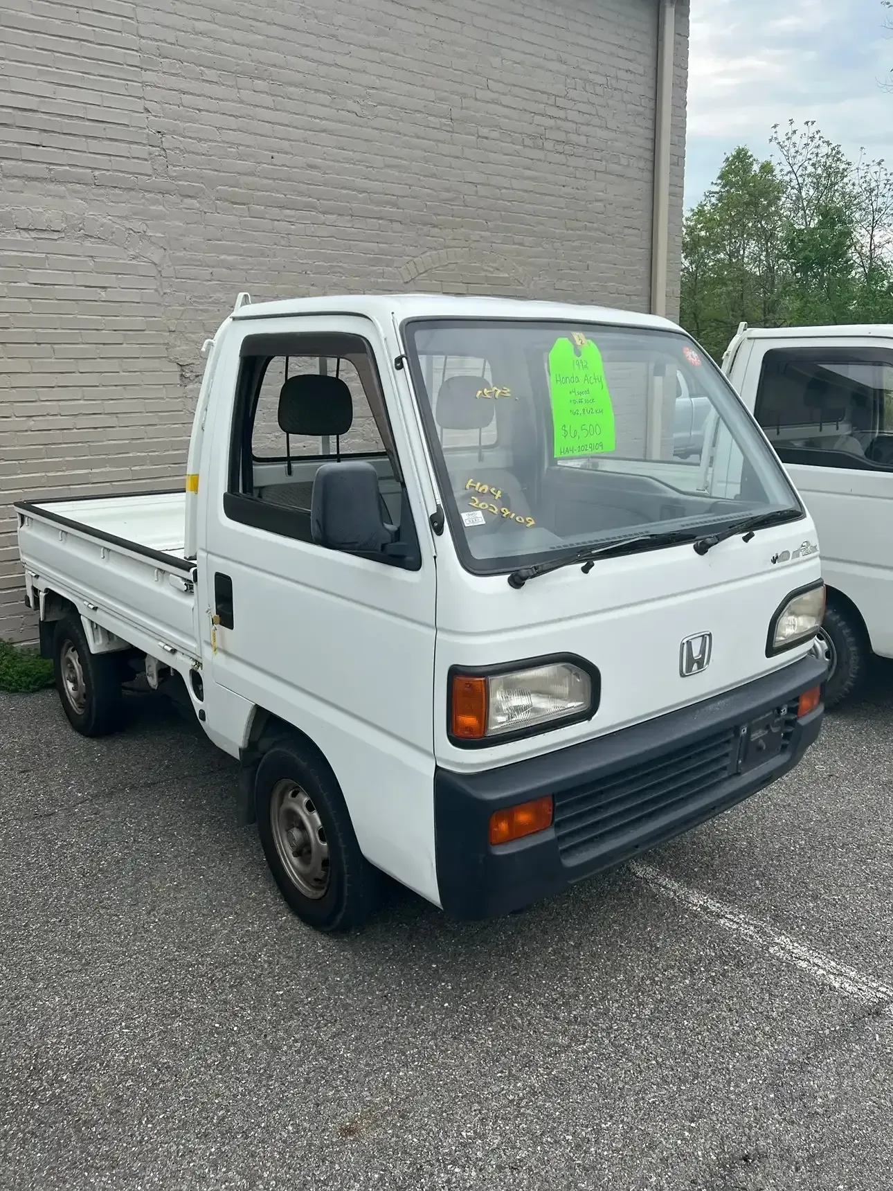 A white, compact Japanese kei truck parked on an asphalt lot in front of a brick building.