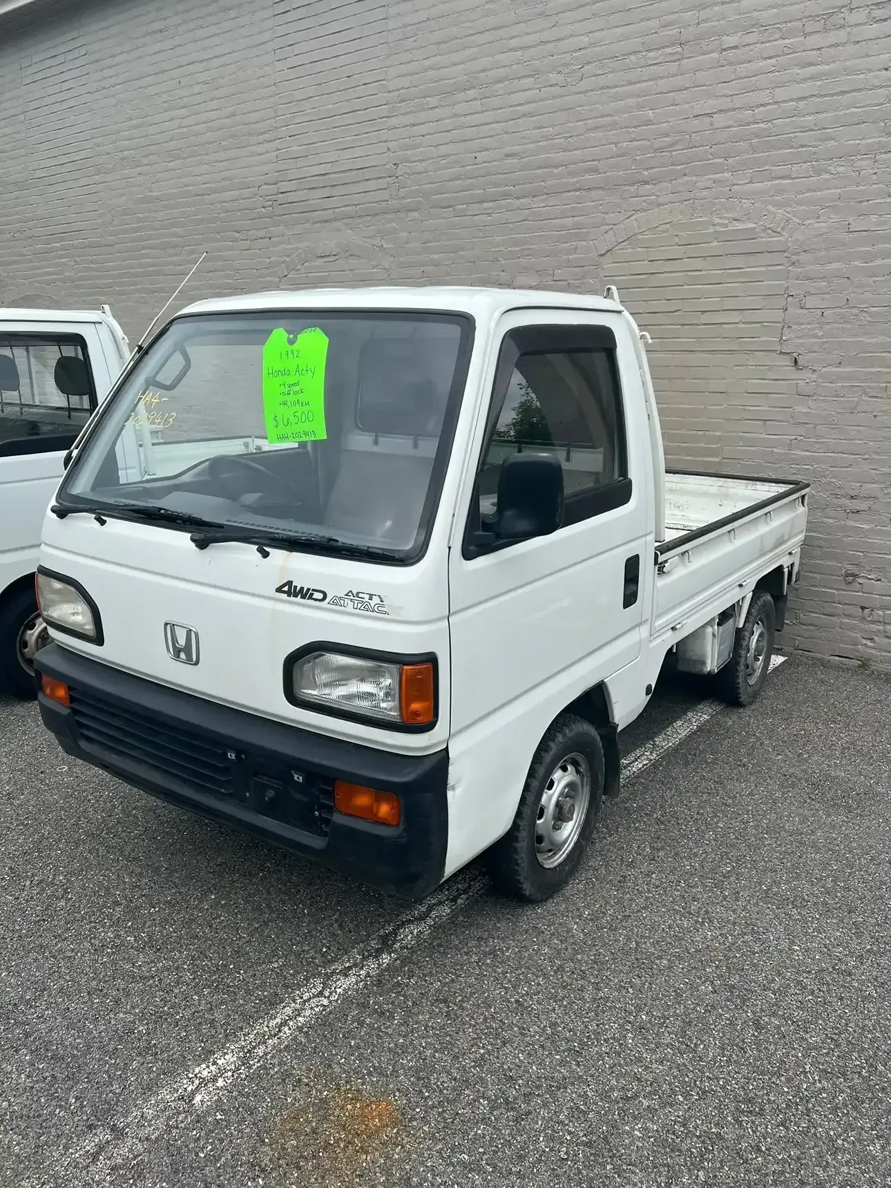 A white Honda Acty mini truck parked on an asphalt lot in front of a gray brick wall.