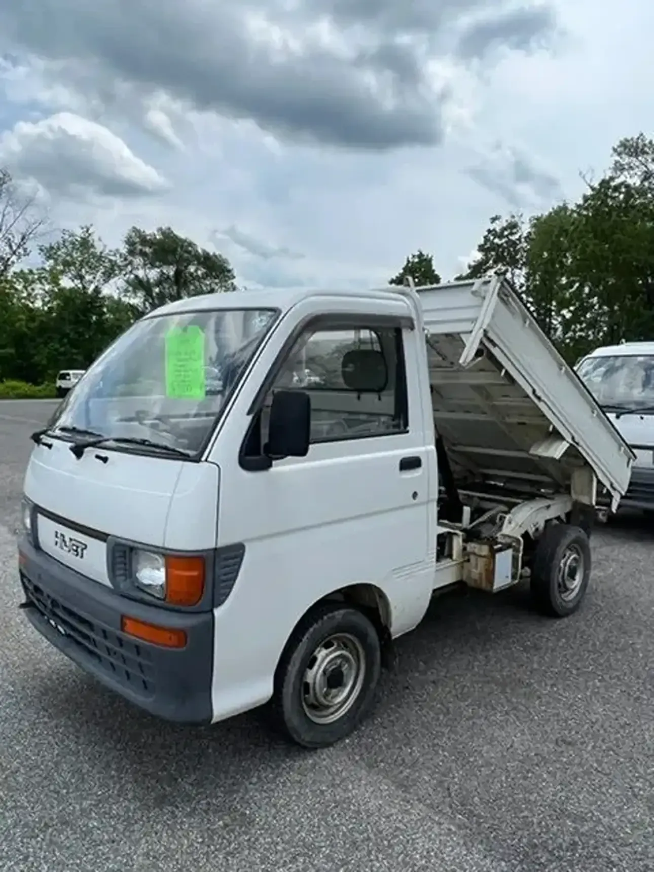 A white, compact dump truck with its bed raised, parked on an asphalt lot under a cloudy sky.