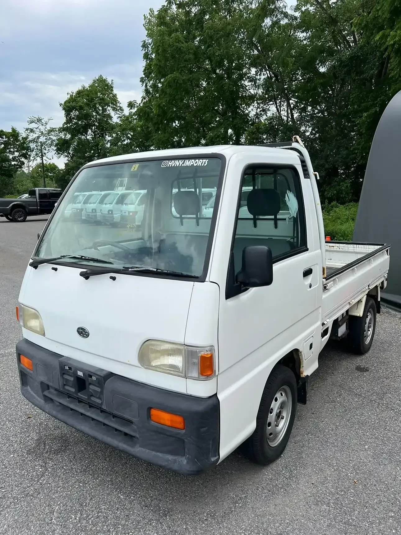 A white, right-hand drive kei truck parked on a gravel lot with trees in the background.